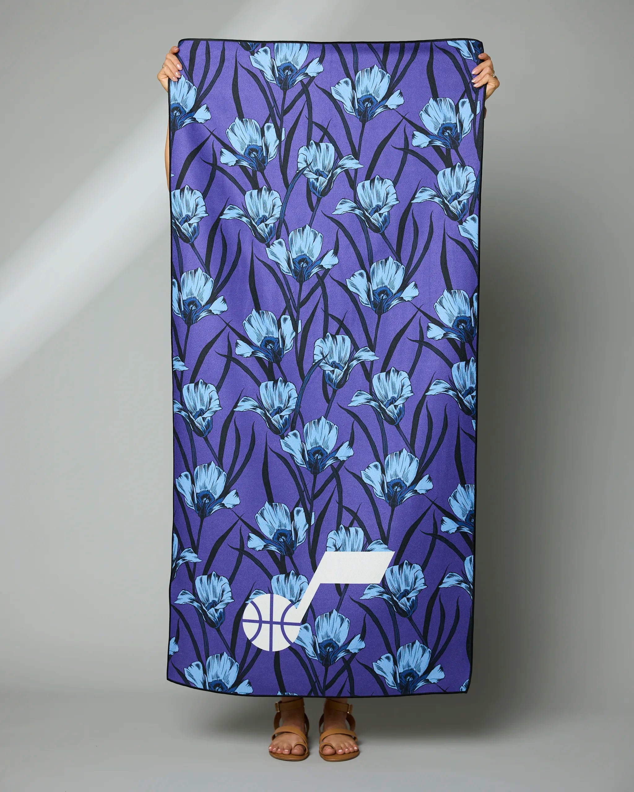 A person holds the Geometry Jazz Floral towel, featuring a blue floral pattern on a dark background and a white geometric logo in the corner; only their hands, feet, and sandals are visible below the towel.