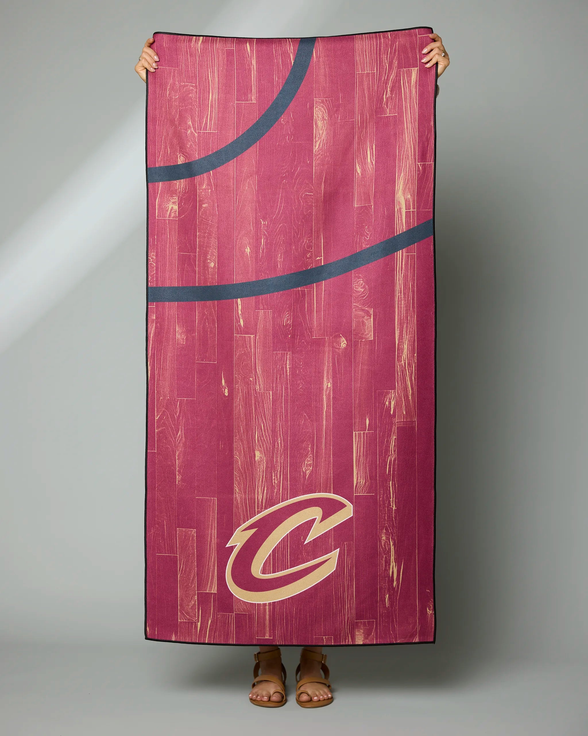 A person holds up the Geometry Cavaliers Zone towel, featuring a maroon woodgrain basketball court design with black lines and a gold Cleveland Cavaliers C logo in the corner. Only their hands and feet are visible.