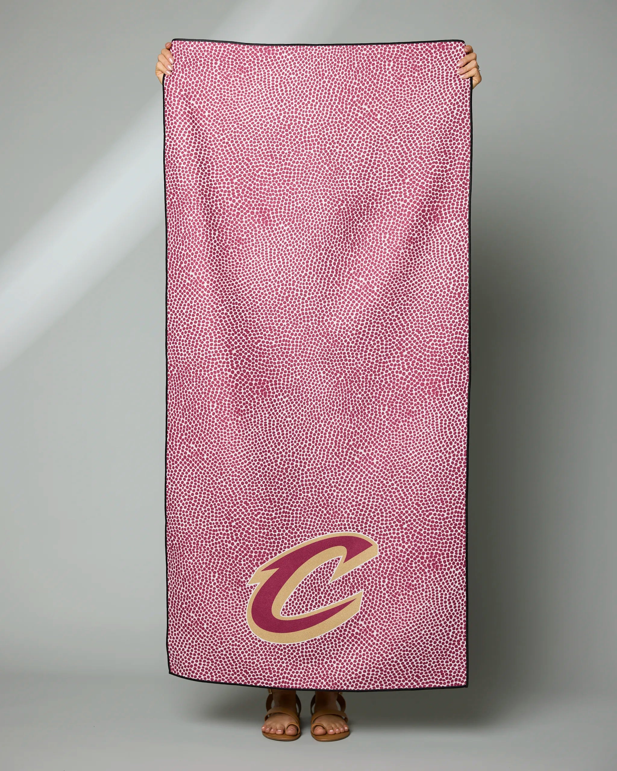 A person holds the Geometry Cavaliers Grip towel, pink with a small white dot pattern and a large yellow and red “C” logo in the bottom right corner. Their hands and feet are visible, but their face is hidden behind the towel.