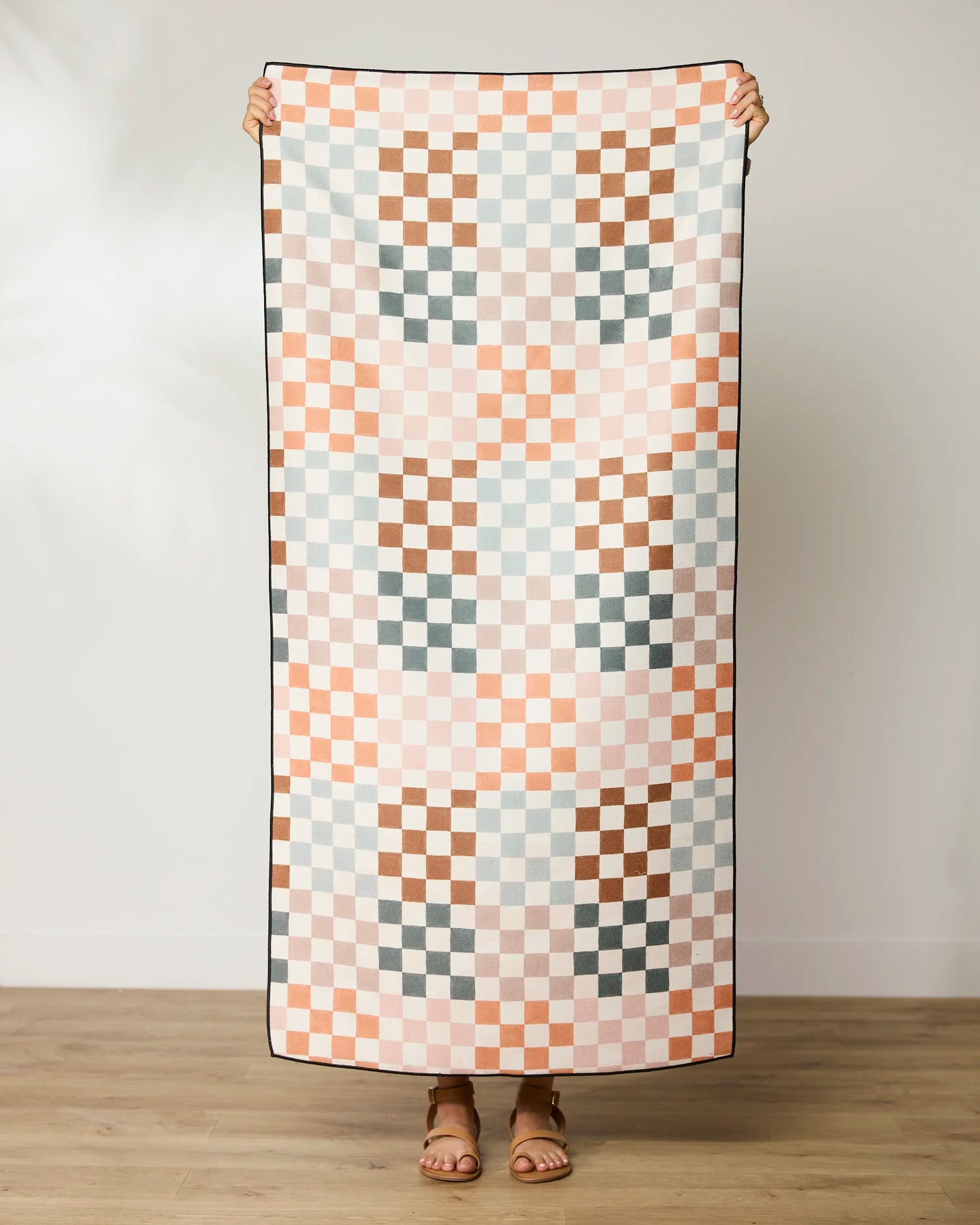 A person stands holding up the GEOMETRY Autumn Checkers micro-loop beach towel, featuring a pastel pink, peach, brown, and light blue checkerboard pattern. Their head is not visible and they wear sandals on a wooden floor.