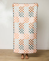 A person stands holding up the GEOMETRY Autumn Checkers micro-loop beach towel, featuring a pastel pink, peach, brown, and light blue checkerboard pattern. Their head is not visible and they wear sandals on a wooden floor.