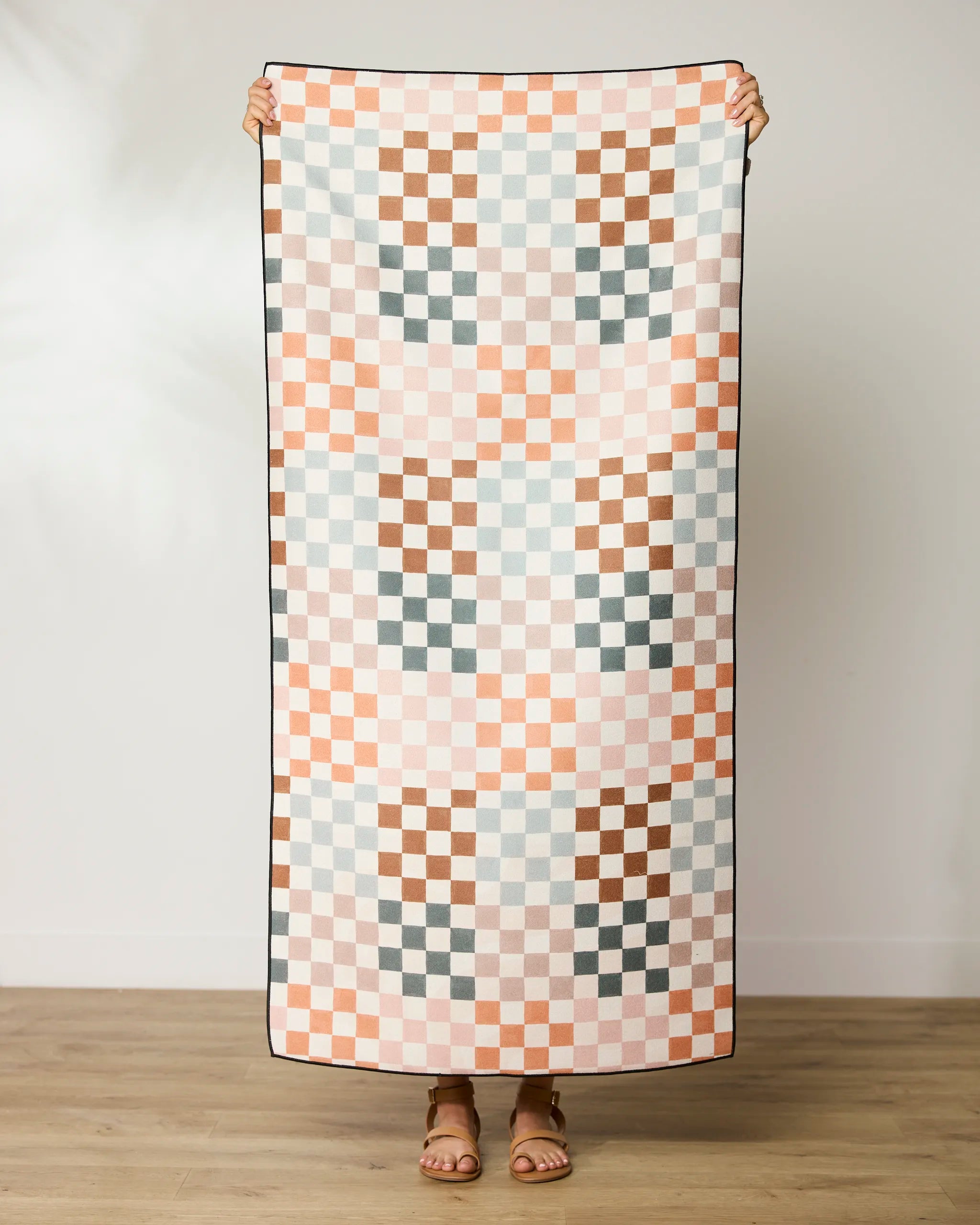 A person stands holding up the GEOMETRY Autumn Checkers micro-loop beach towel, featuring a pastel pink, peach, brown, and light blue checkerboard pattern. Their head is not visible and they wear sandals on a wooden floor.