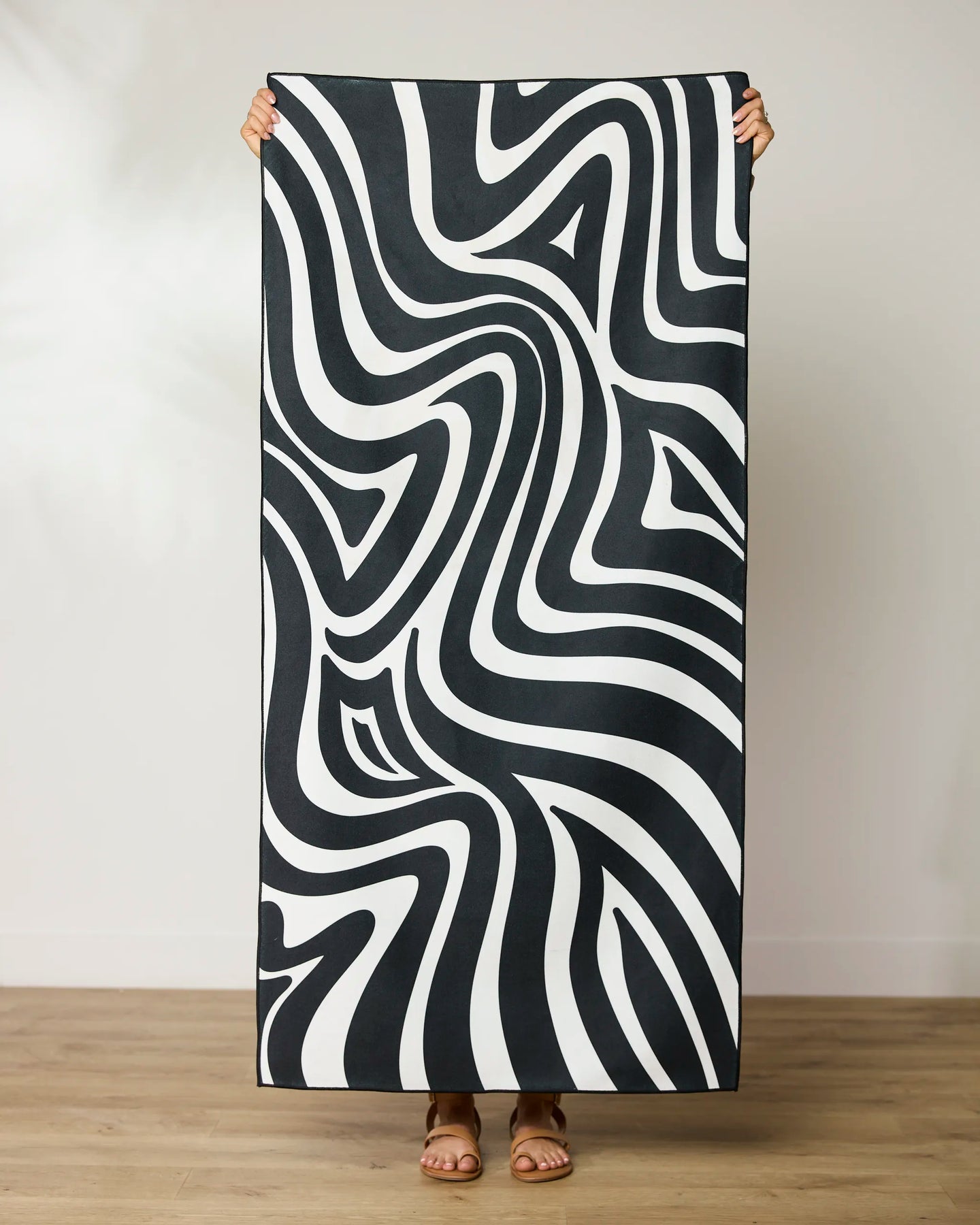 A person stands on a wooden floor, holding up the GEOMETRY Dizzy Black quick-drying beach towel featuring bold black and white wavy abstract patterns that create an optical illusion. Their face and upper body are hidden behind the towel.