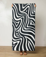 A person stands on a wooden floor, holding up the GEOMETRY Dizzy Black quick-drying beach towel featuring bold black and white wavy abstract patterns that create an optical illusion. Their face and upper body are hidden behind the towel.