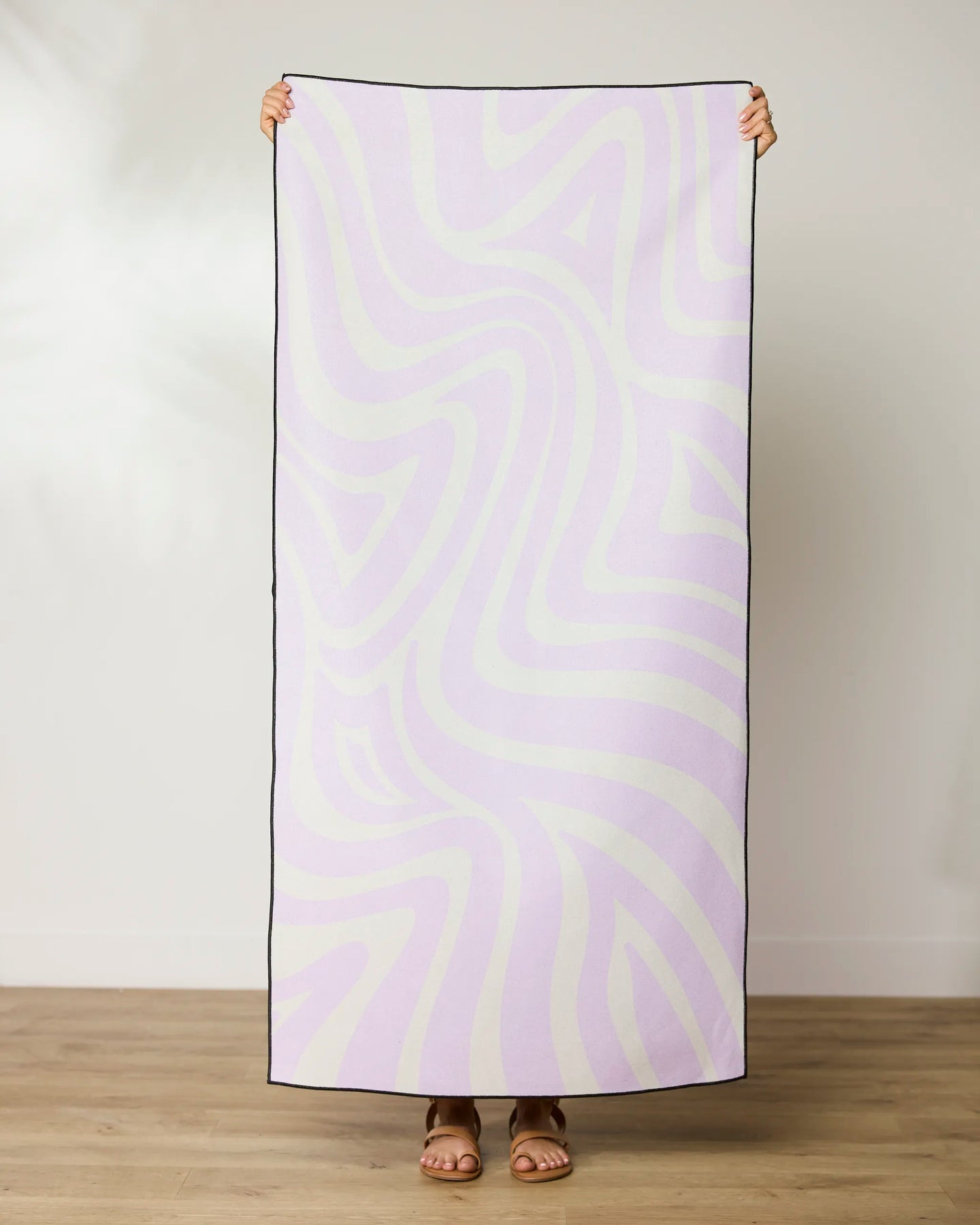 A person stands on a wooden floor, holding up the GEOMETRY Dizzy Lilac towel—made from recycled materials with a pastel purple and white wavy design. Only their hands and sandaled feet are seen behind the towel.