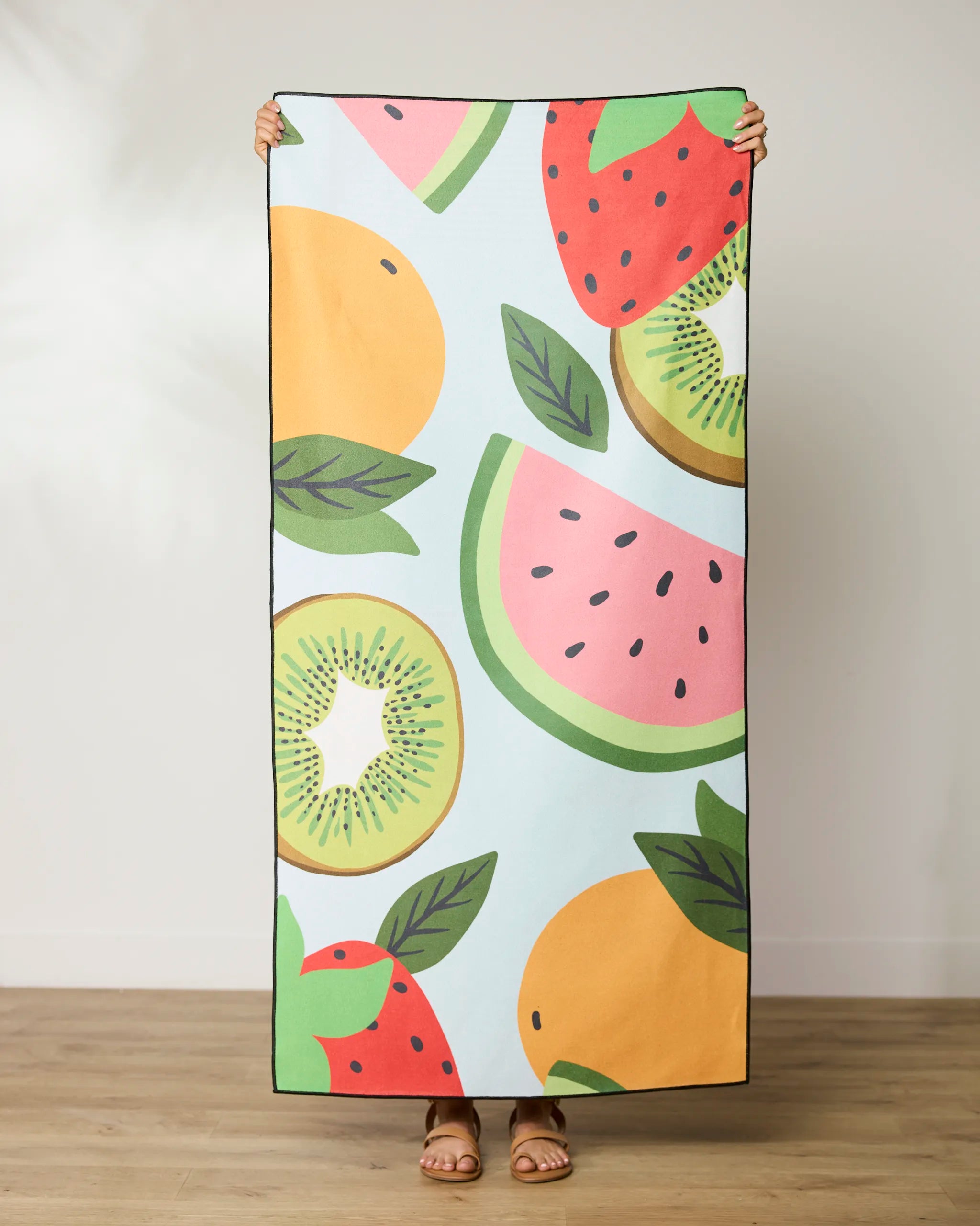 A person stands on a wooden floor holding up the vibrant GEOMETRY Fruit Salad Micro-loop Beach Towel, featuring oranges, kiwis, strawberries, and watermelon slices on a light blue background—made from recycled, quick-drying materials.