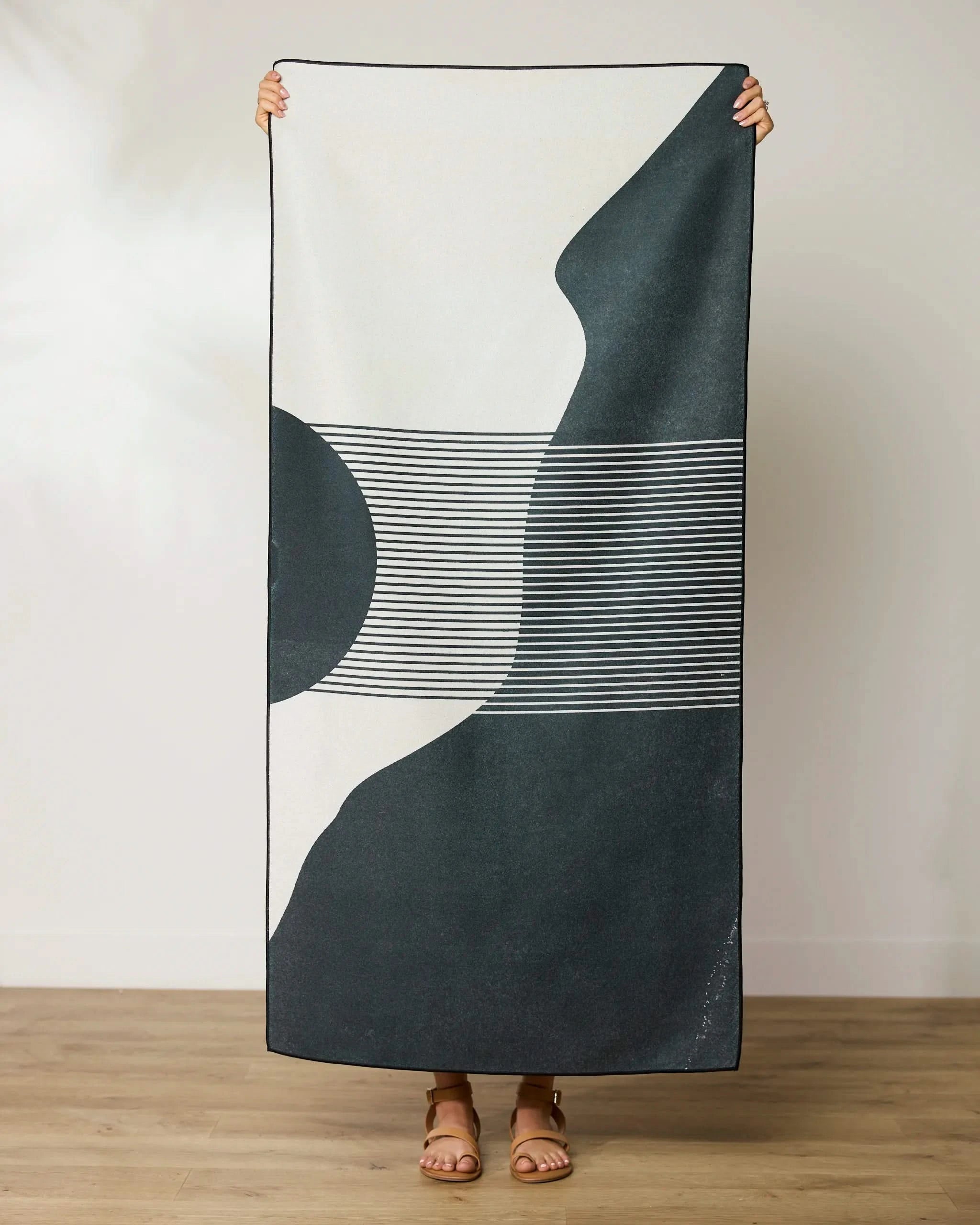 Only their sandaled feet show as someone stands on a wooden floor, holding up the GEOMETRY Geo Hill Beach Towel—crafted from recycled materials and featuring bold black-and-white abstract geometric patterns.