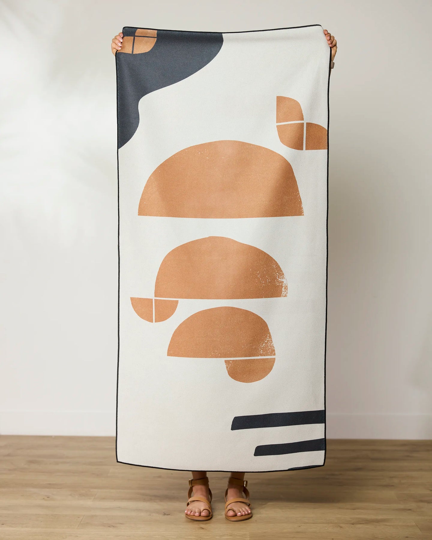 A person stands indoors holding the GEOMETRY Geo Phases quick-drying beach towel, featuring modern abstract shapes in tan, black, and white. Only their hands, feet, and sandals are visible behind the towel.