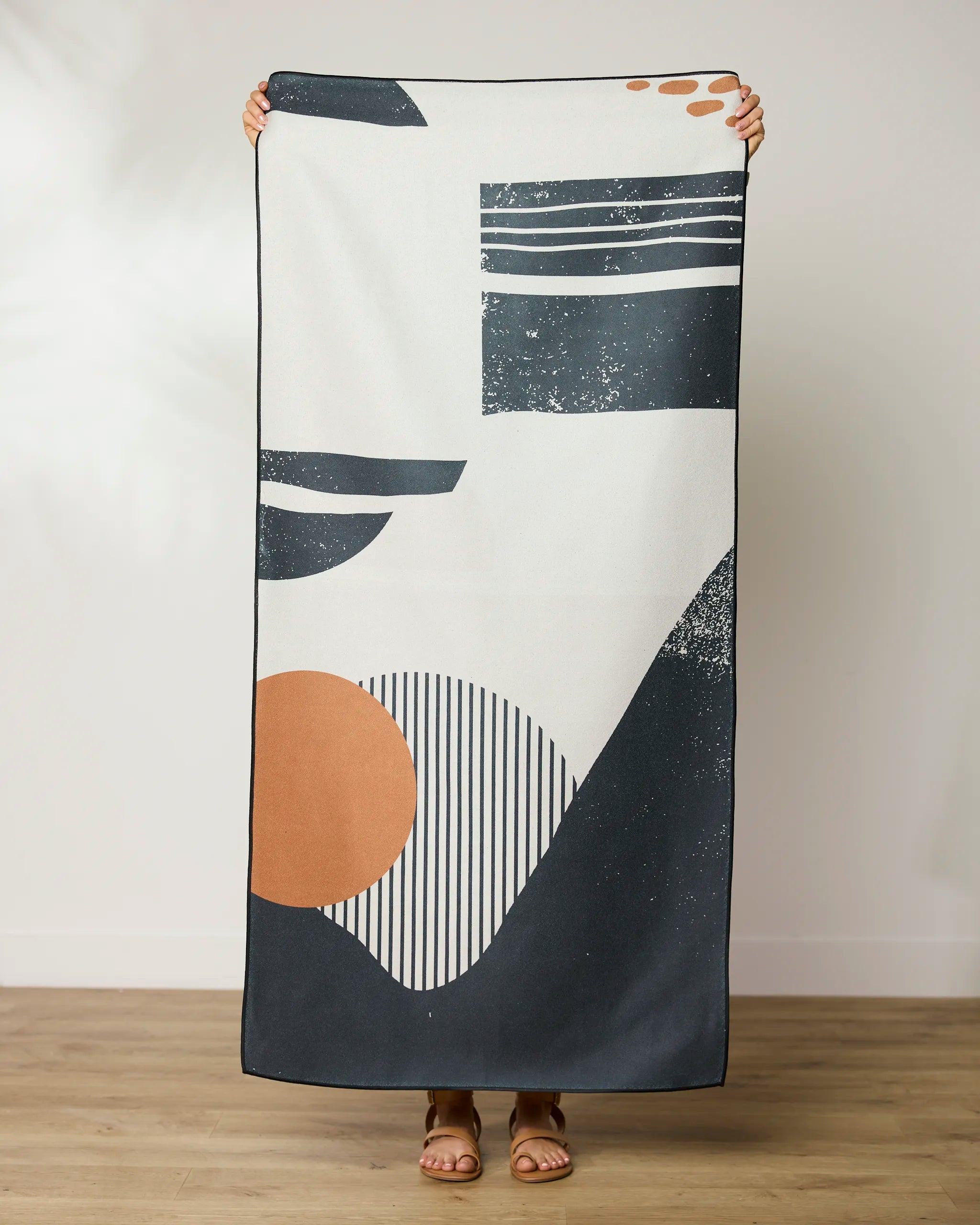 A person stands barefoot on a wooden floor, holding up a GEOMETRY Geo Seaside Beach Towel featuring a modern abstract design with black, white, and rust-colored geometric shapes and lines.