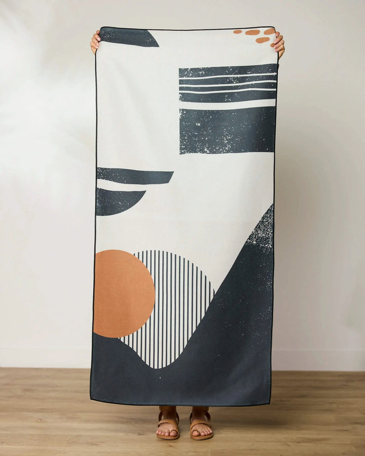 A person stands barefoot on a wooden floor, holding up a GEOMETRY Geo Seaside Beach Towel featuring a modern abstract design with black, white, and rust-colored geometric shapes and lines.