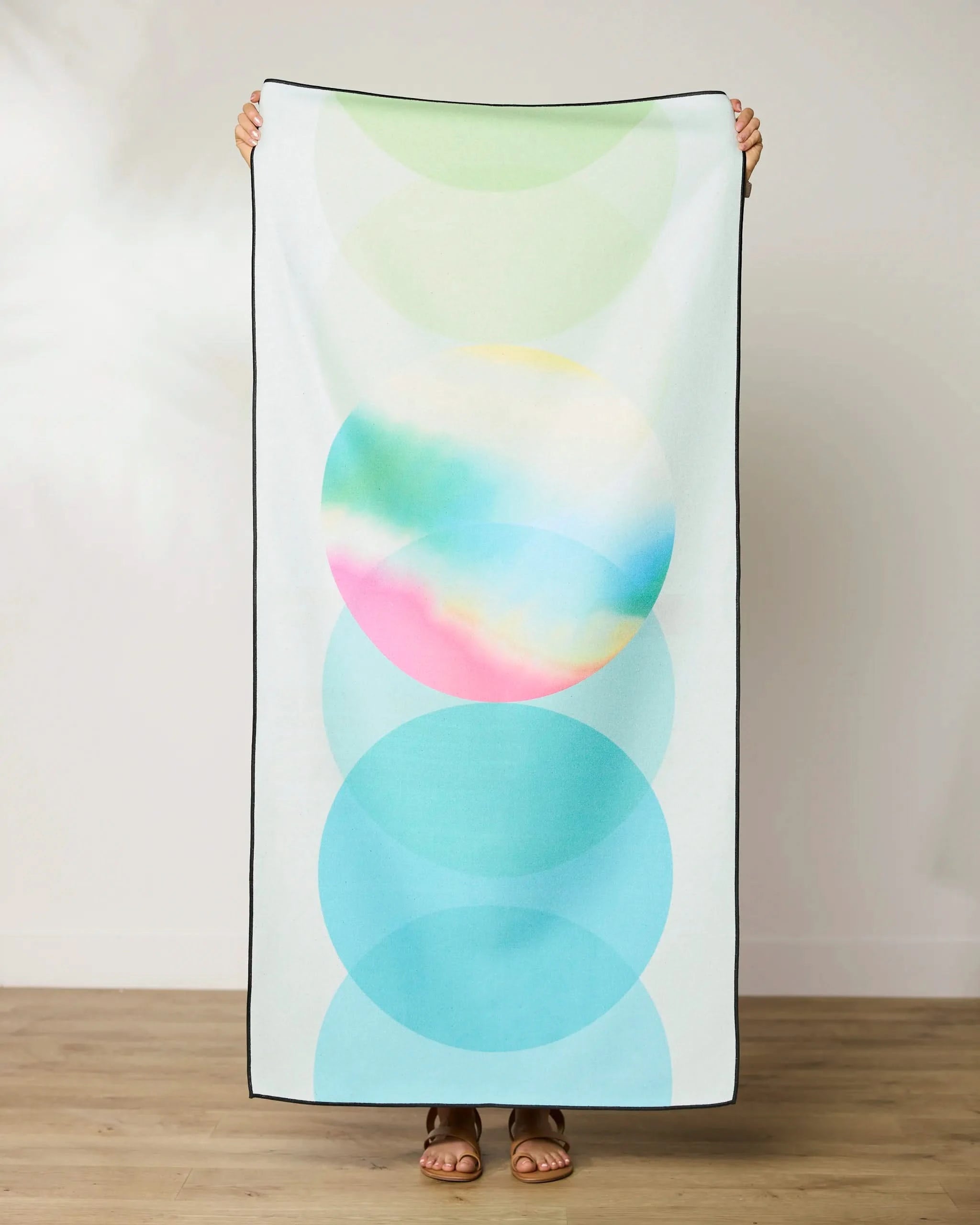 A person indoors holds up the GEOMETRY Green Dream quick-drying micro-loop beach towel, showcasing overlapping pastel circles in green, yellow, pink, and blue on a white background. Only their hands and feet are visible.