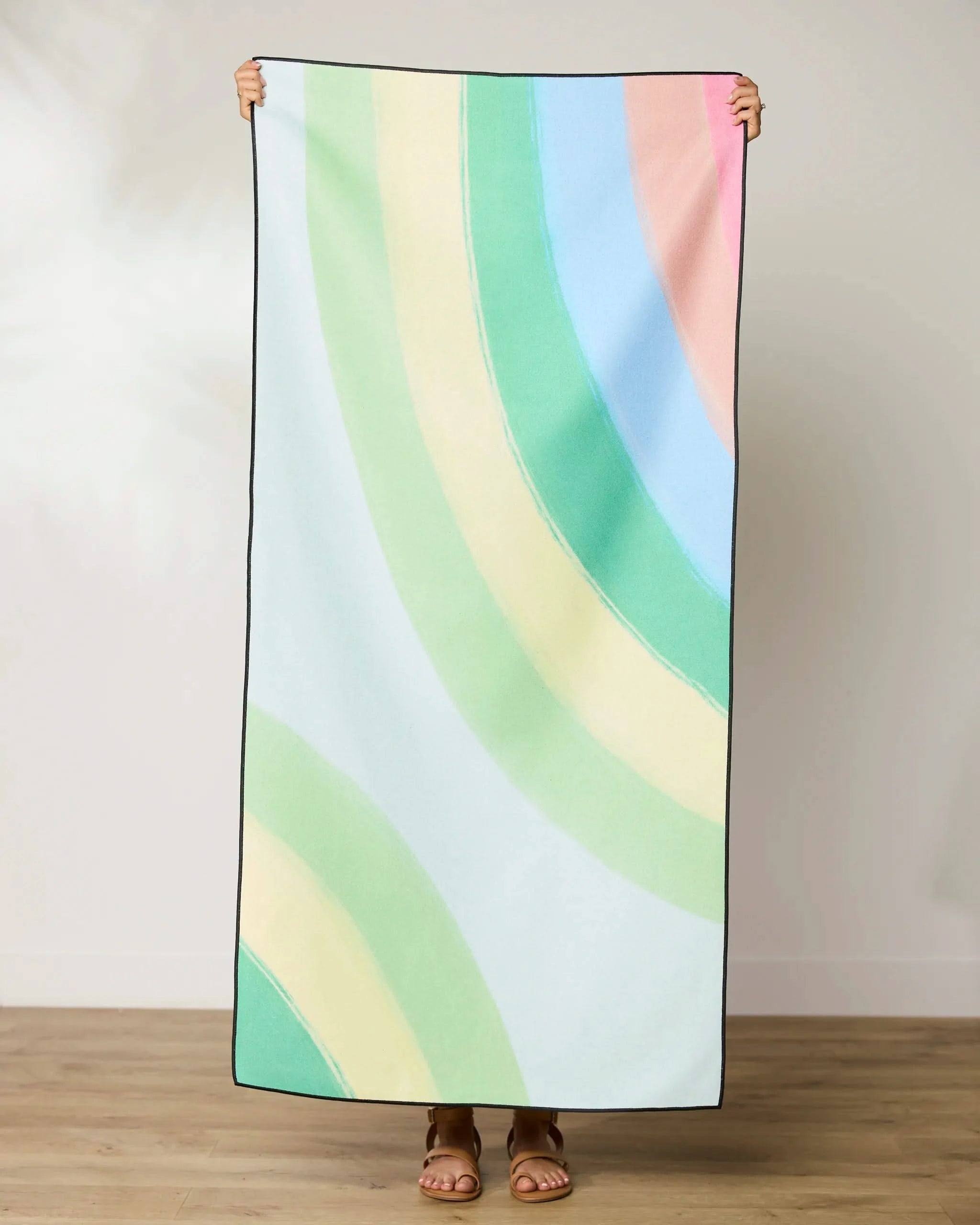 Indoors on a wooden floor, a person holds up the GEOMETRY Green Rainbow beach towel featuring pastel green, yellow, blue, and pink arcs. The quick-drying towel conceals their face—only their feet are visible.