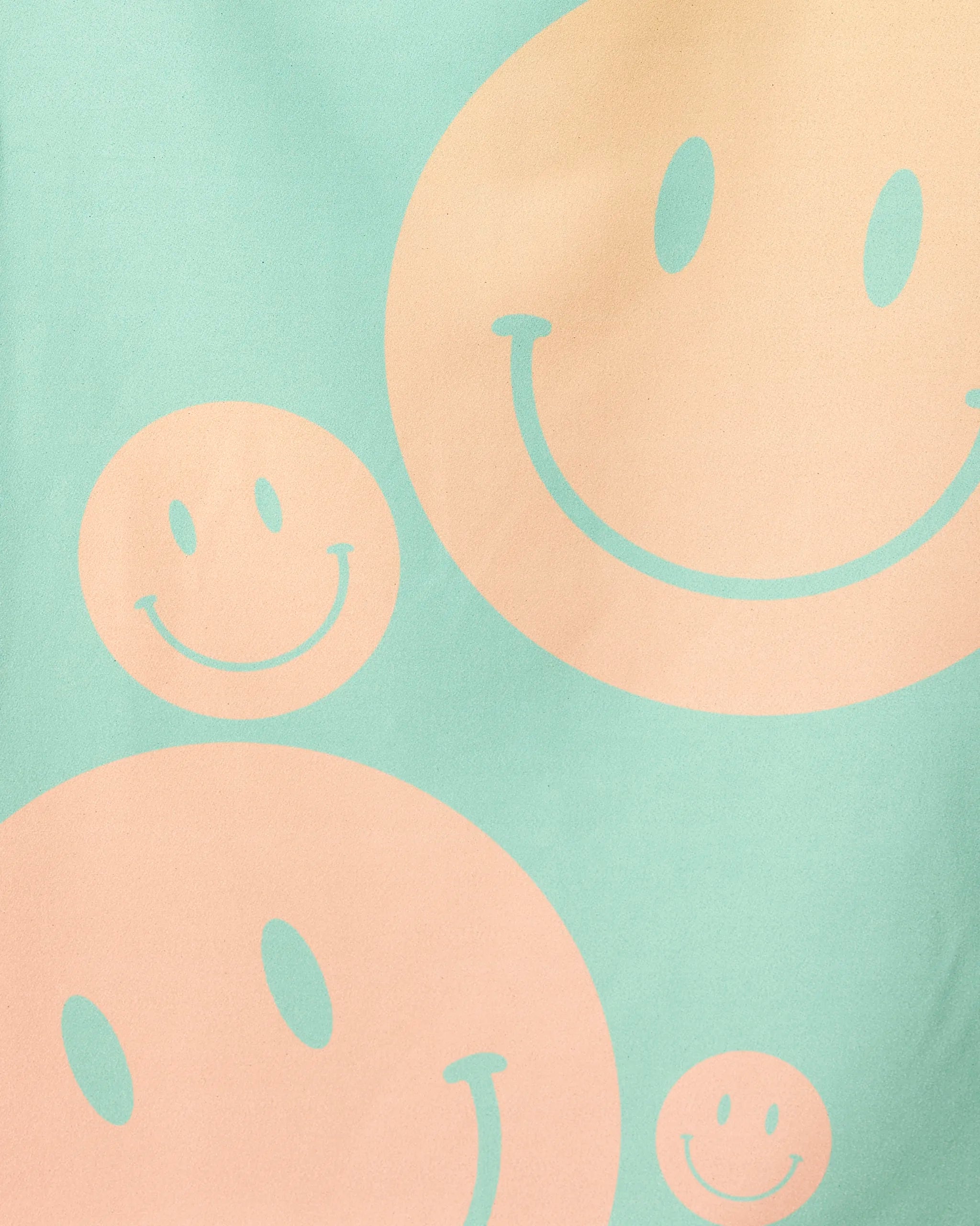 A light green background displays large and small peach-colored smiley faces with curved smiles and oval eyes, inspired by the playful design of GEOMETRY’s Happy Days Micro-loop Beach Towel made from recycled materials.