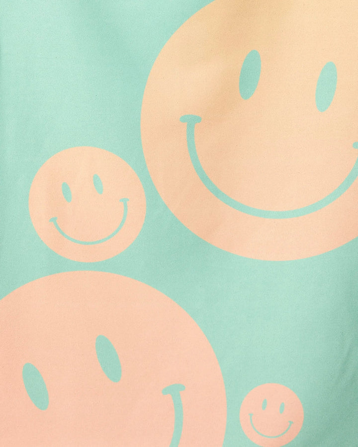 A light green background displays large and small peach-colored smiley faces with curved smiles and oval eyes, inspired by the playful design of GEOMETRY’s Happy Days Micro-loop Beach Towel made from recycled materials.