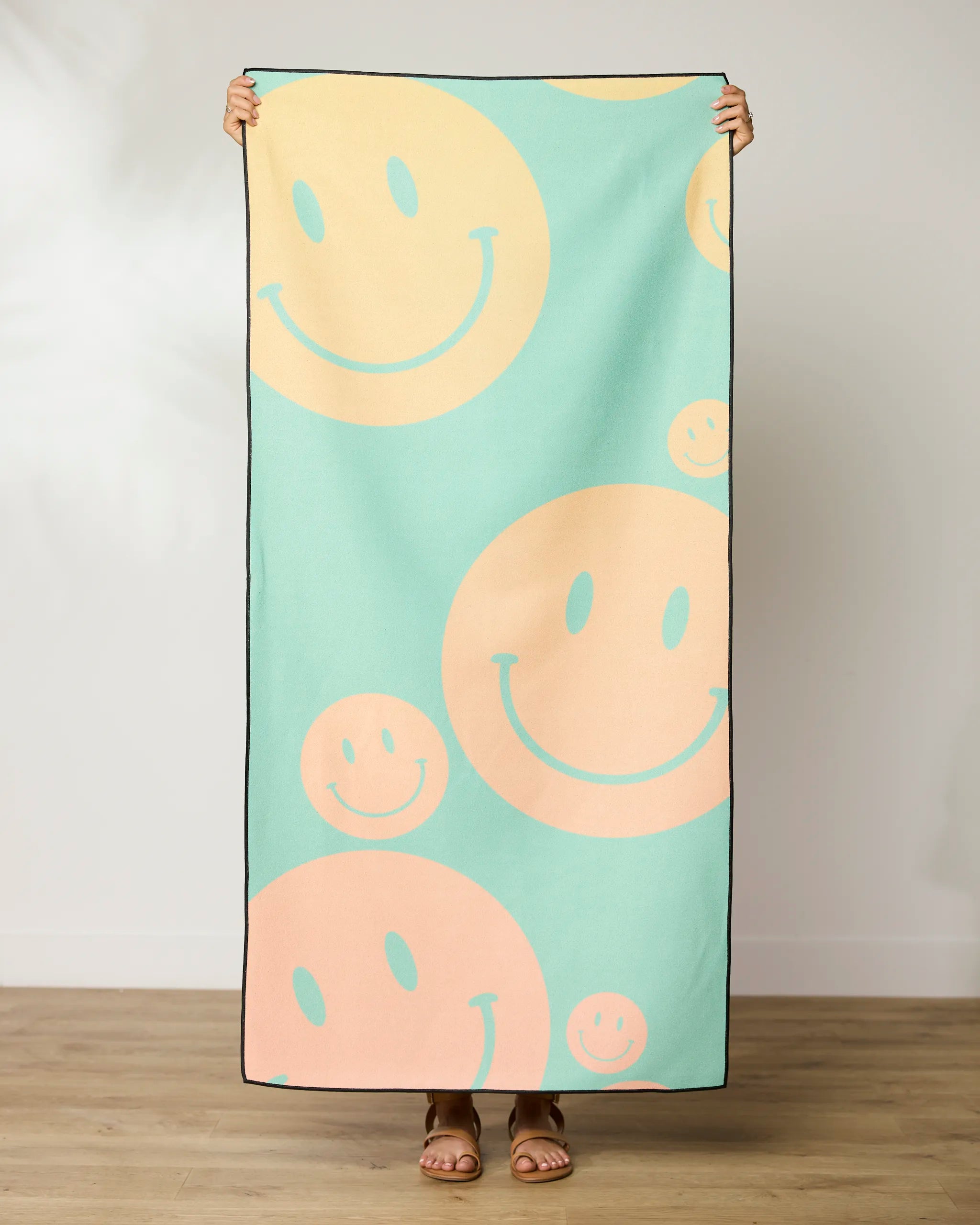 A person stands indoors holding up the GEOMETRY Happy Days Micro-loop Beach Towel, featuring a pastel green background with large peach and yellow smiley faces. Only their hands and feet are visible. Made from recycled materials.