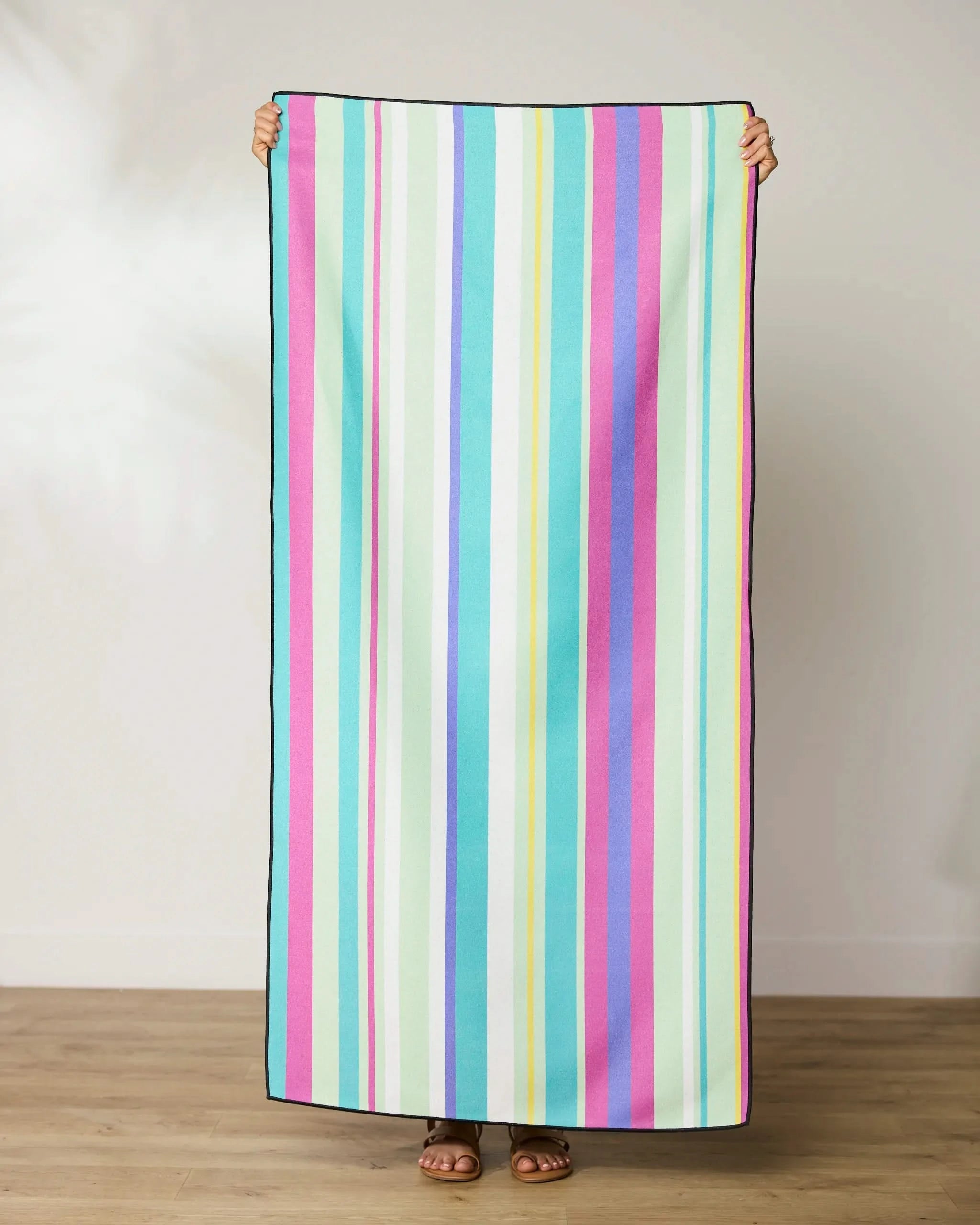 A person stands on a wooden floor, holding up the GEOMETRY Line It Up Grape Beach Towel with vertical pastel stripes, covering their body except for feet and hands.