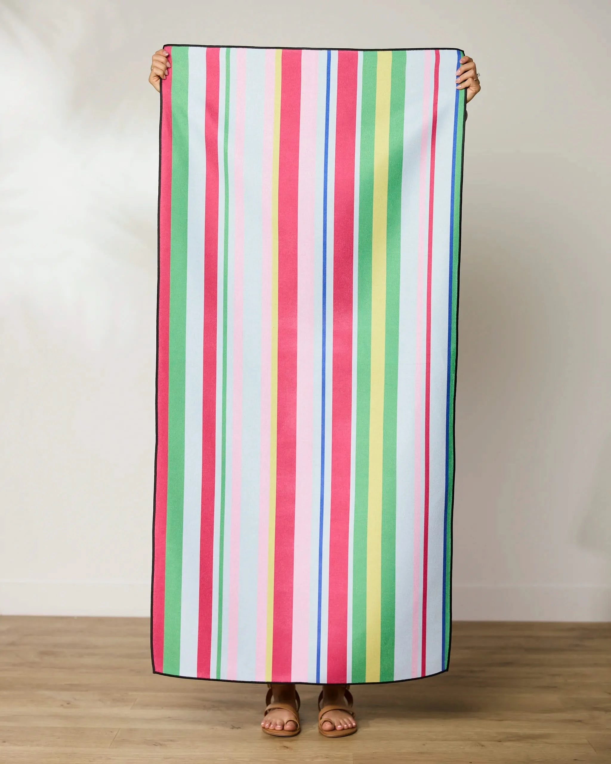 Indoors, a person holds up the GEOMETRY Line It Up Melon Micro-loop Beach Towel with vertical pink, red, blue, green, and white stripes, covering their body from shoulders to feet; only hands and feet are visible.