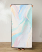 A person holds up GEOMETRYs Mellow Micro-loop Beach Towel, showcasing its quick-drying recycled material and soft pastel abstract pattern in blue, pink, and cream. Only their hands and sandaled feet are visible.