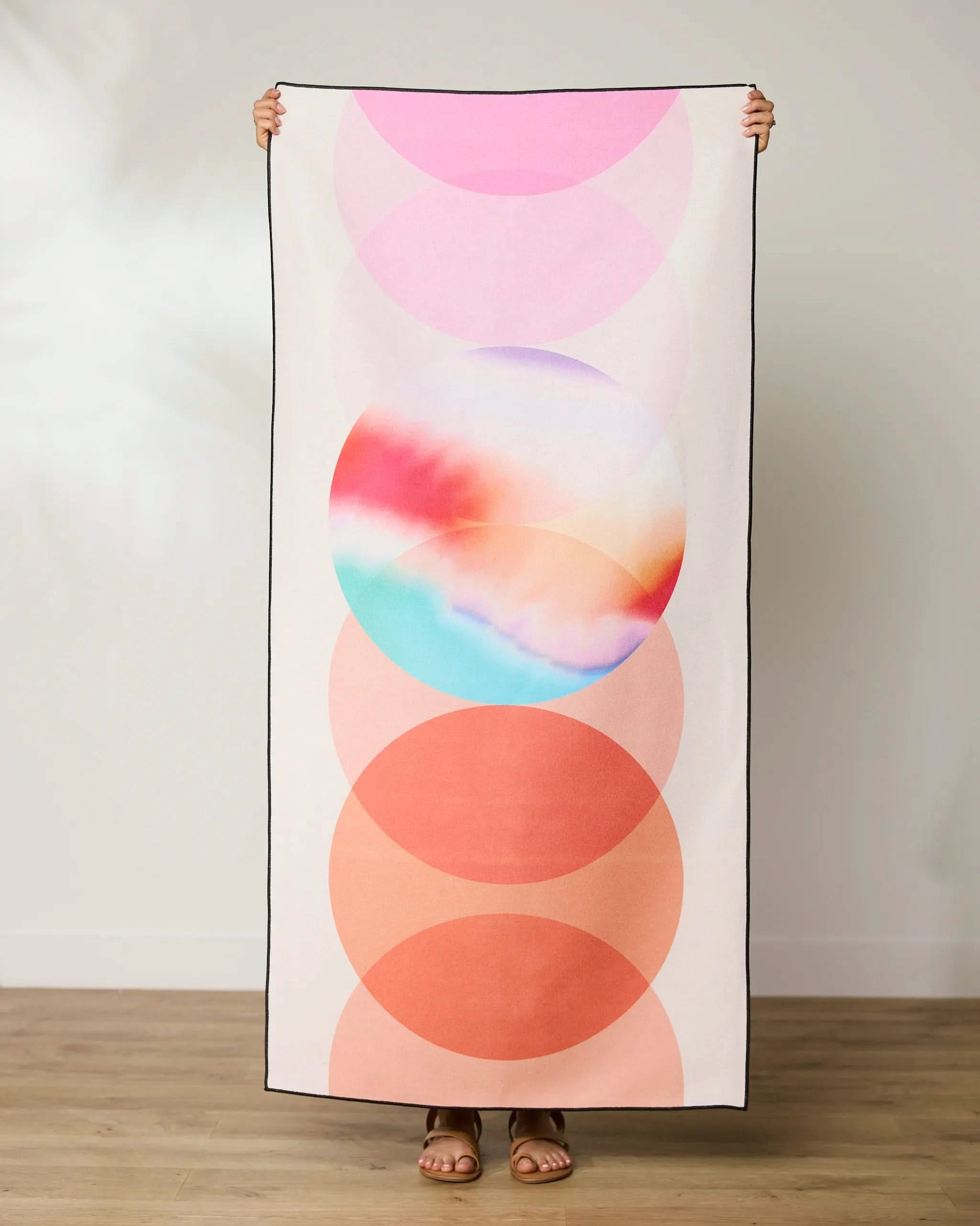 A person holds up the GEOMETRY Orange Dream Beach Towel with abstract circular designs in pink, orange, and a multicolored center. Only their hands and feet are visible against a plain wall and wooden floor.