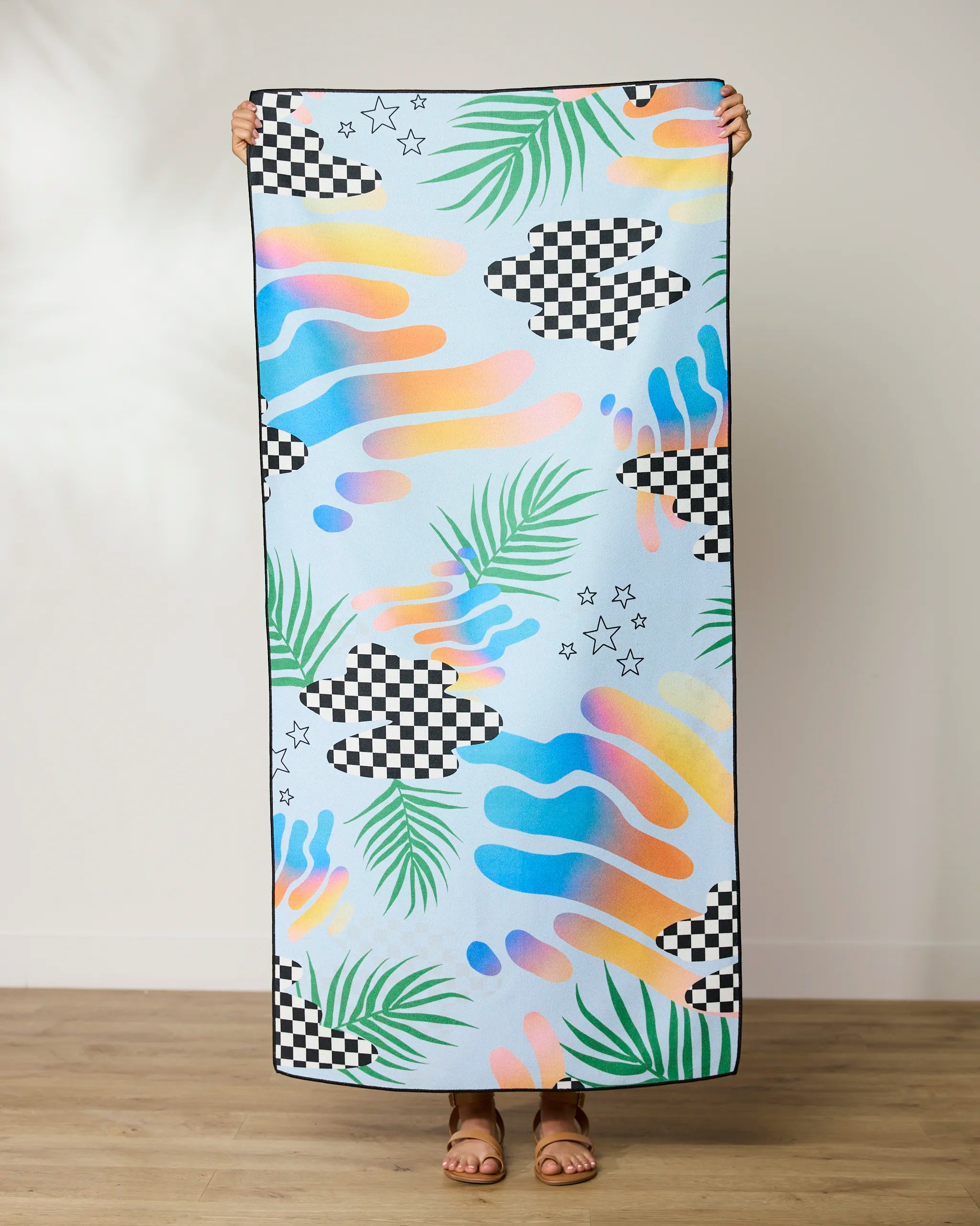 Someone holds up the GEOMETRY Playful Paradise Beach Towel, featuring colorful checkerboard patterns, pastel swirls, green palm leaves, and black stars—a vibrant, quick-drying towel made from recycled materials.