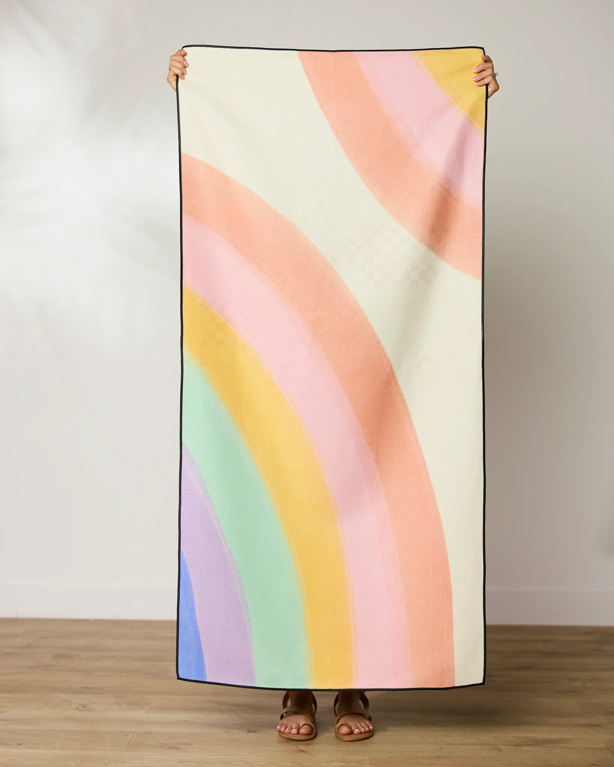 A person stands on a wooden floor holding up a GEOMETRY Rainbow Micro-loop Beach Towel with pastel rainbow stripes on cream; only their feet and hands are visible.