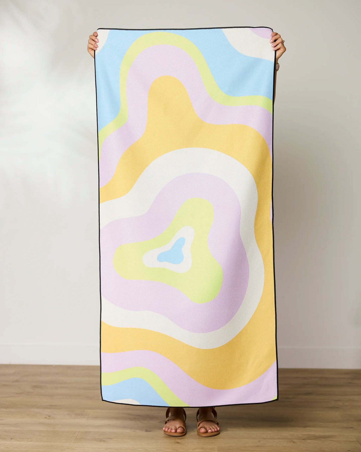 A person stands on a wooden floor holding up a GEOMETRY Swirls Beach Towel with abstract pastel shapes in yellow, purple, blue, green, and white. Their head and hands appear above this quick-drying towel.