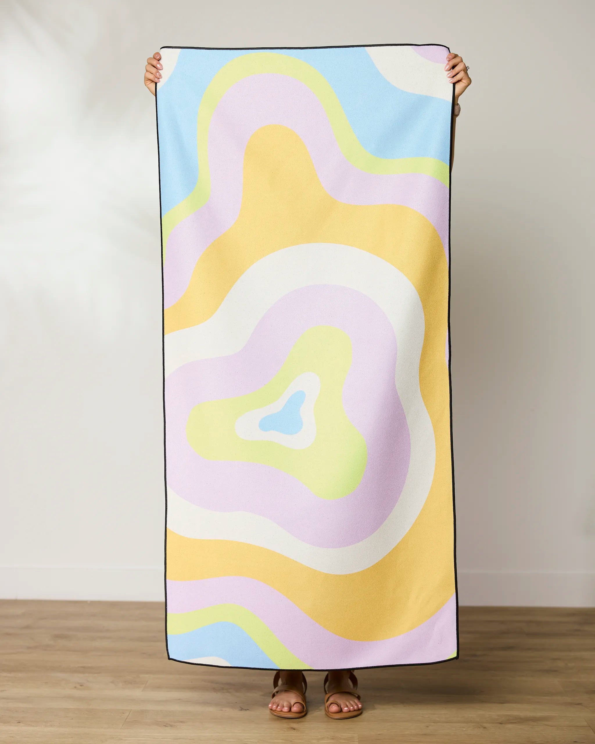 A person stands on a wooden floor holding up a GEOMETRY Swirls Beach Towel with abstract pastel shapes in yellow, purple, blue, green, and white. Their head and hands appear above this quick-drying towel.