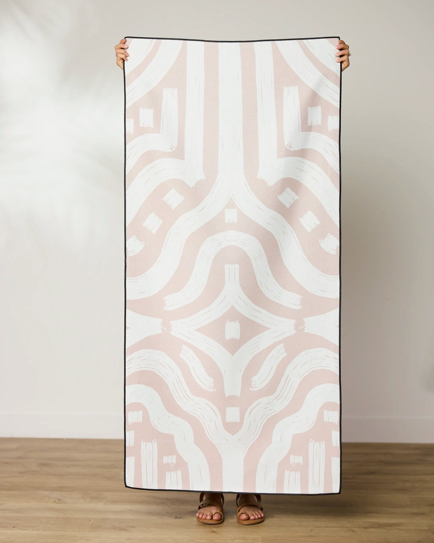 A person stands holding a large GEOMETRY Tribal Beach Towel featuring an abstract geometric pattern in soft pink and white, with black trimmed edges. The towel covers their face and upper body. The floor beneath is wooden.