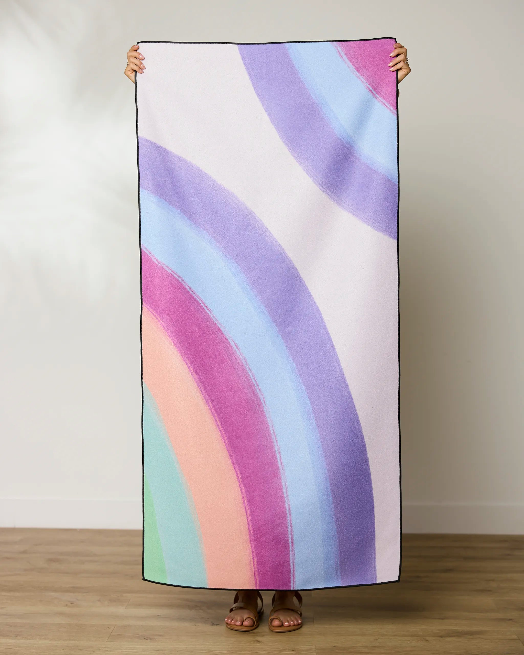 A person stands on a wooden floor, holding up the GEOMETRY Violet Rainbow Micro-loop Beach Towel made from recycled materials. Their head is hidden behind the quick-drying towel, set against a plain light wall.