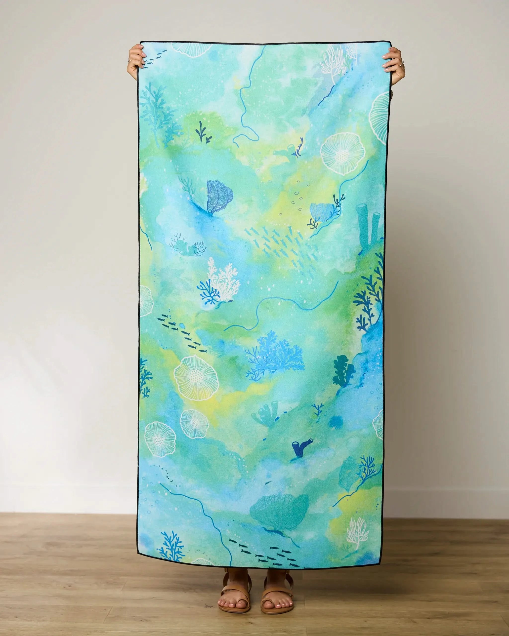 A person stands indoors holding up the GEOMETRY Reefy Greens quick-drying beach towel, featuring an aquatic design in blue and green with coral, fish, and sea plants. Only their feet and hands are visible.