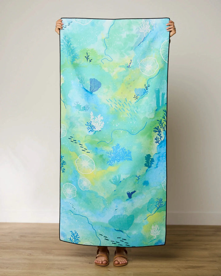 A person stands indoors holding up the GEOMETRY Reefy Greens quick-drying beach towel, featuring an aquatic design in blue and green with coral, fish, and sea plants. Only their feet and hands are visible.