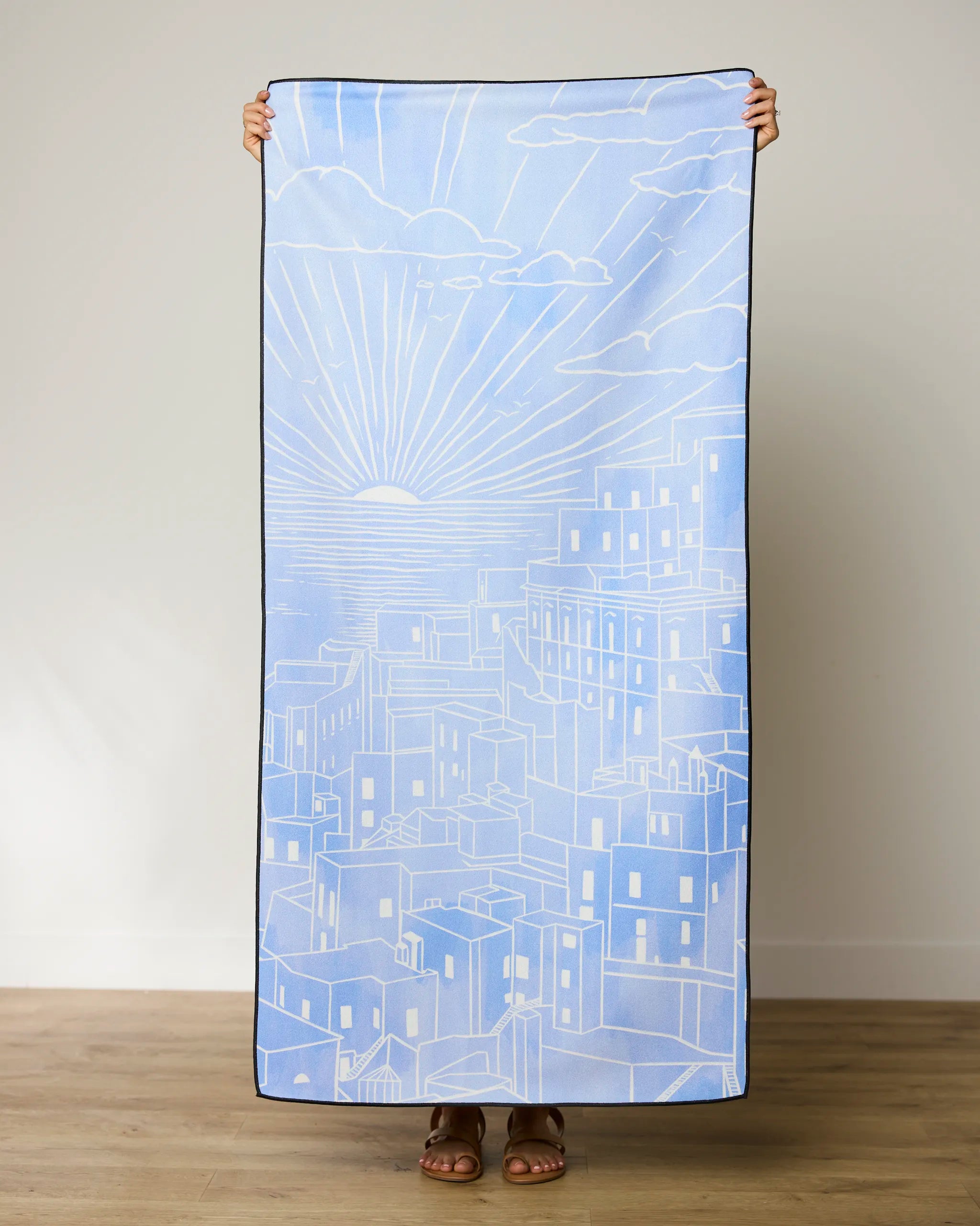 A person stands barefoot on a wooden floor, holding up the GEOMETRY Amalfi Coast Beach Towel with a white line drawing of buildings and a sunrise or sunset. The persons face is hidden behind this quick-drying towel.