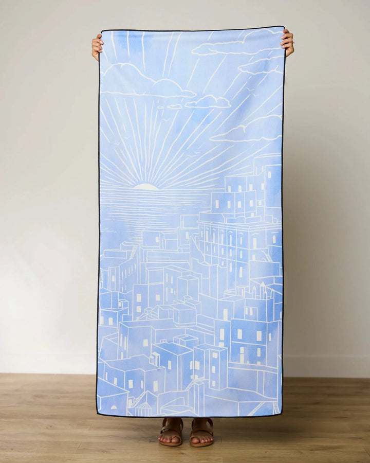 A person stands barefoot on a wooden floor, holding up the GEOMETRY Amalfi Coast Beach Towel with a white line drawing of buildings and a sunrise or sunset. The persons face is hidden behind this quick-drying towel.