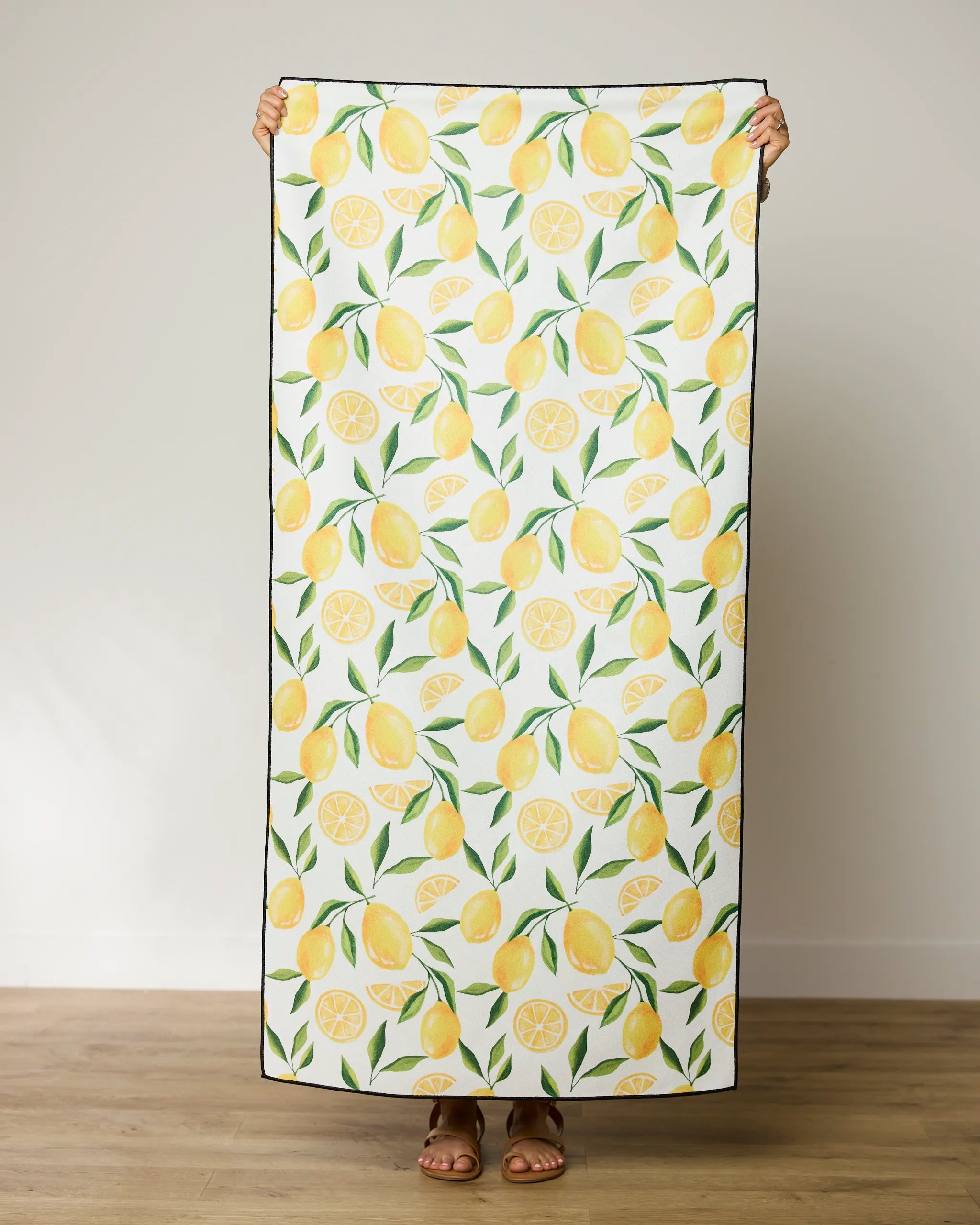 A person holds up the GEOMETRY Limoncello quick-drying micro-loop beach towel—white with whole lemons, slices, and green leaves. Only their hands, feet, and sandals are visible. Made from recycled materials for eco-friendly style.