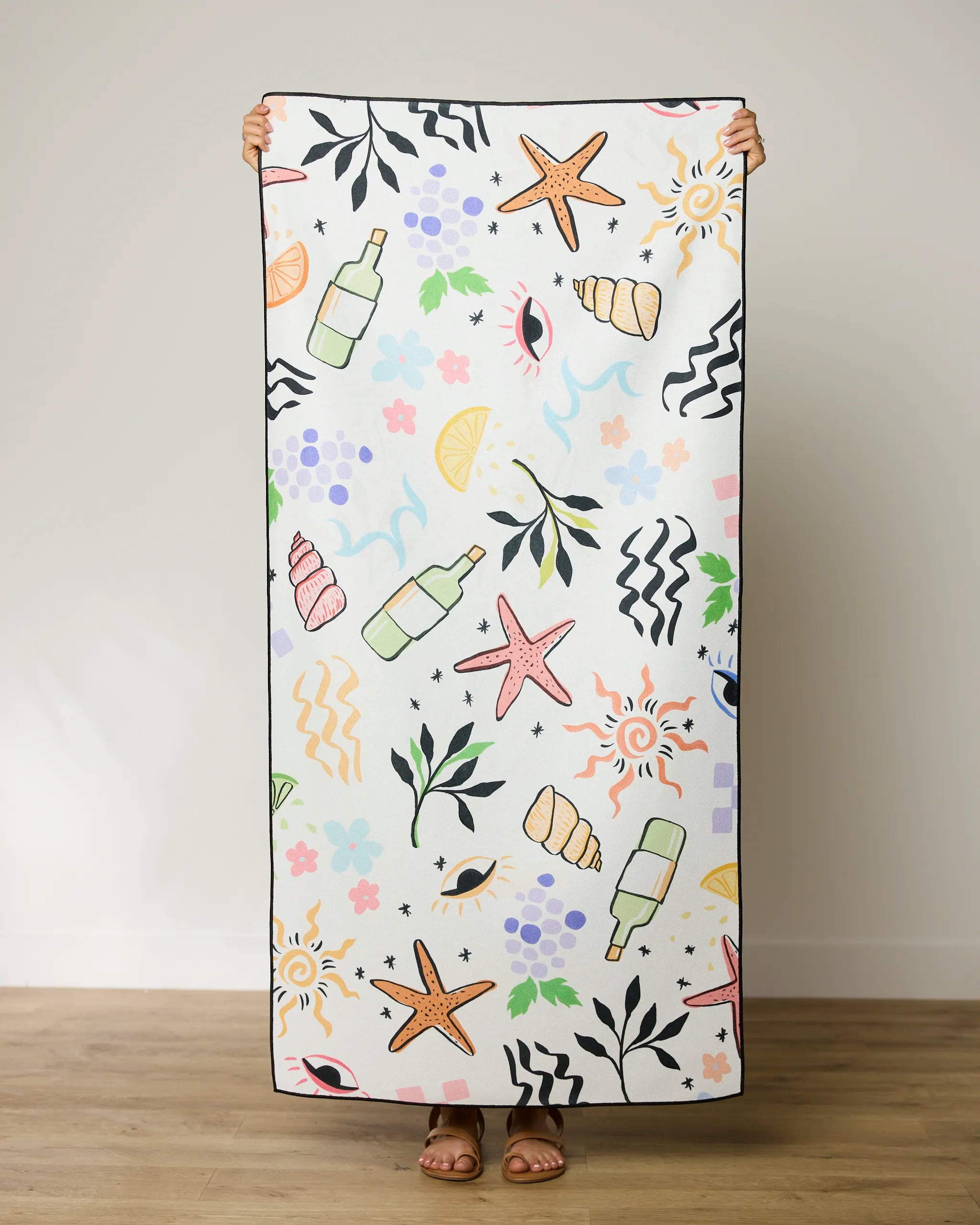 A person stands indoors holding up a GEOMETRY Wine Not beach towel featuring colorful illustrations of starfish, shells, bottles, leaves, fruits, flowers, waves, and abstract shapes. Only the persons feet are visible.