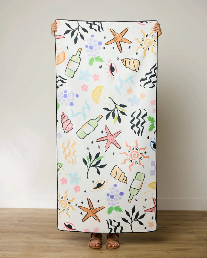A person stands indoors holding up a GEOMETRY Wine Not beach towel featuring colorful illustrations of starfish, shells, bottles, leaves, fruits, flowers, waves, and abstract shapes. Only the persons feet are visible.