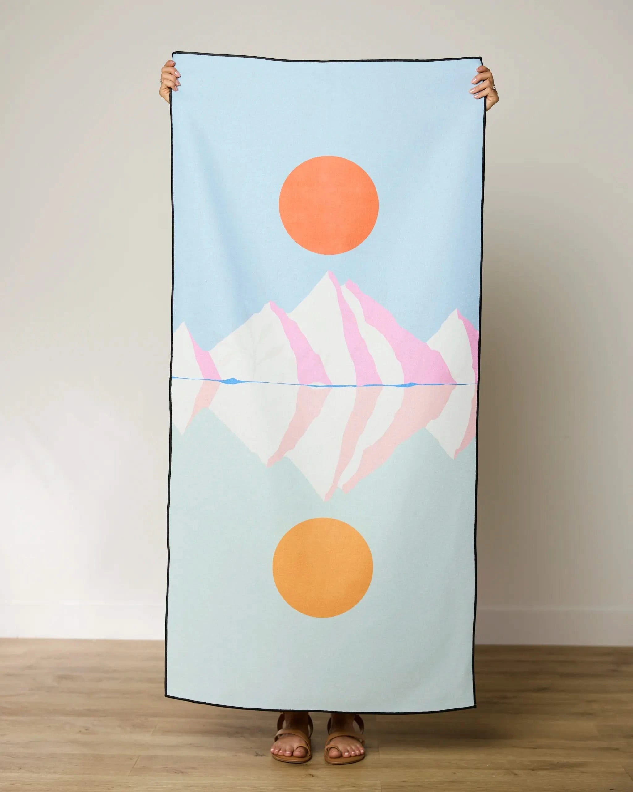 A person holds up the GEOMETRY Elevated Micro-loop Beach Towel, featuring a minimalist design of orange suns and pink mountains on light blue. Only their feet and hands are visible. This quick-drying towel is perfect for sunny days.