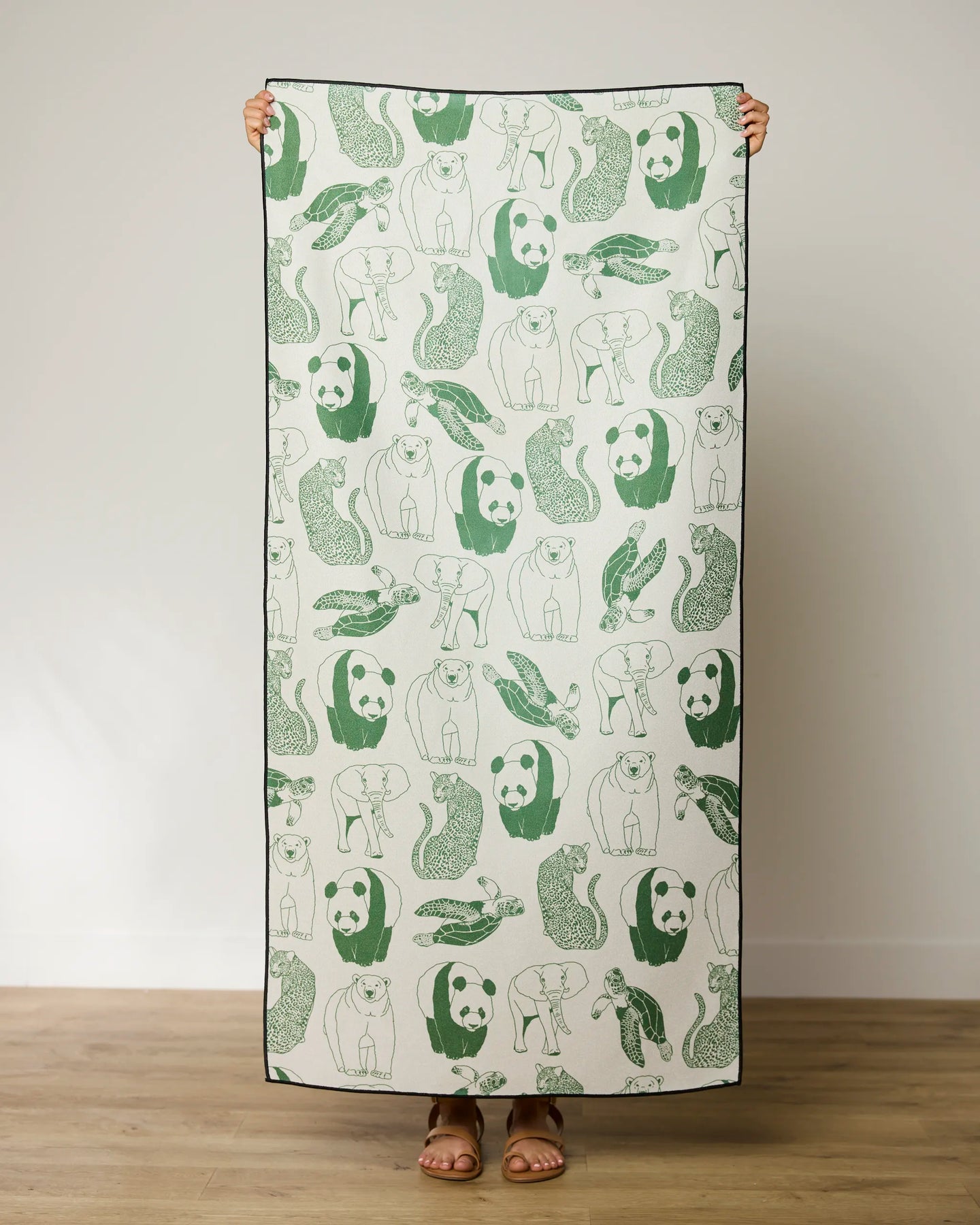 A person stands indoors holding up the GEOMETRY Endangered micro-loop beach towel, featuring green and gray line drawings of pandas, bears, otters, and rabbits. Only their sandal-clad feet are visible below the quick-drying towel.