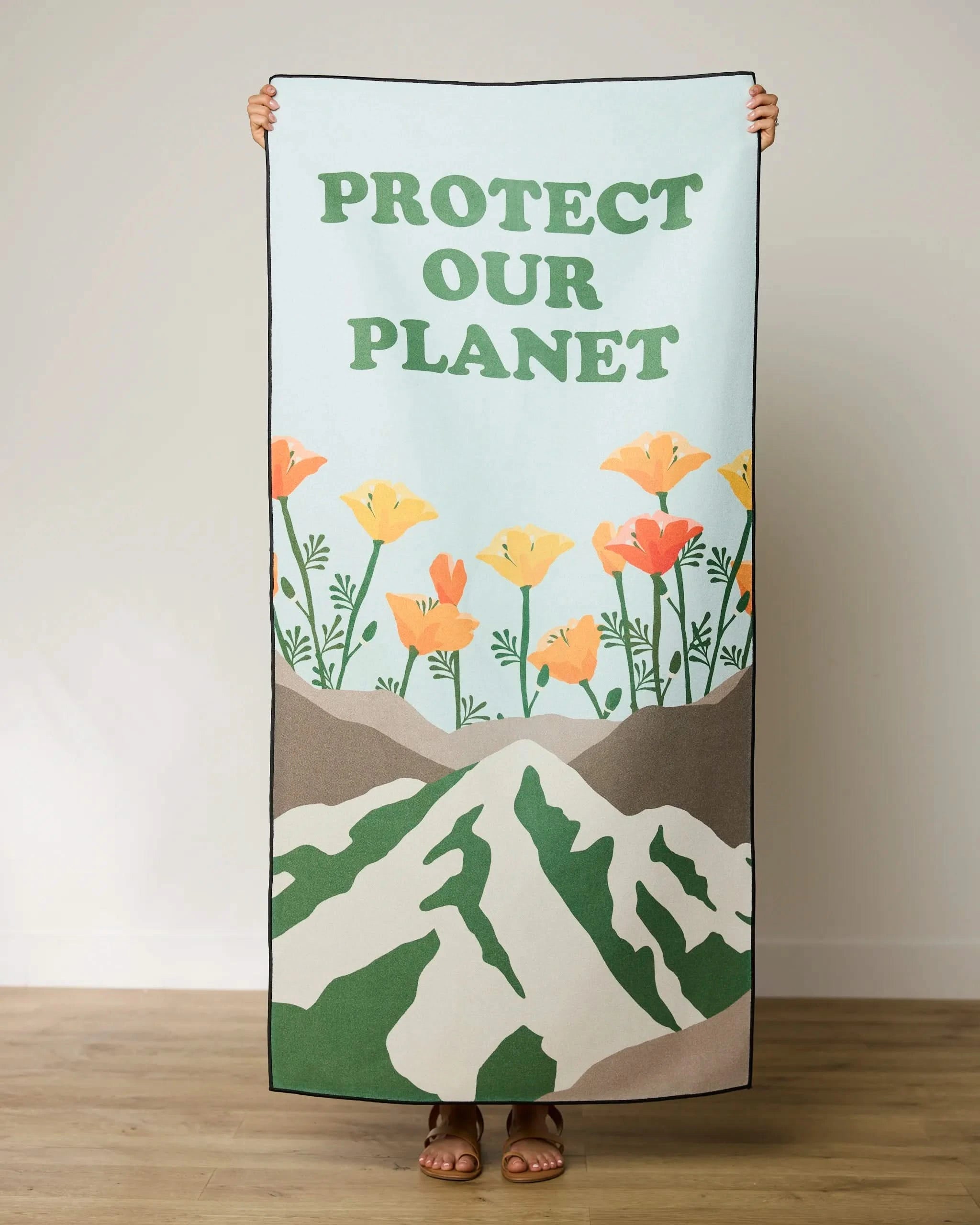 A person holds up GEOMETRY’s Protect beach towel, which features orange flowers, green mountains, and PROTECT OUR PLANET in bold green letters. Only their hands and feet are visible—made from quick-drying recycled materials.