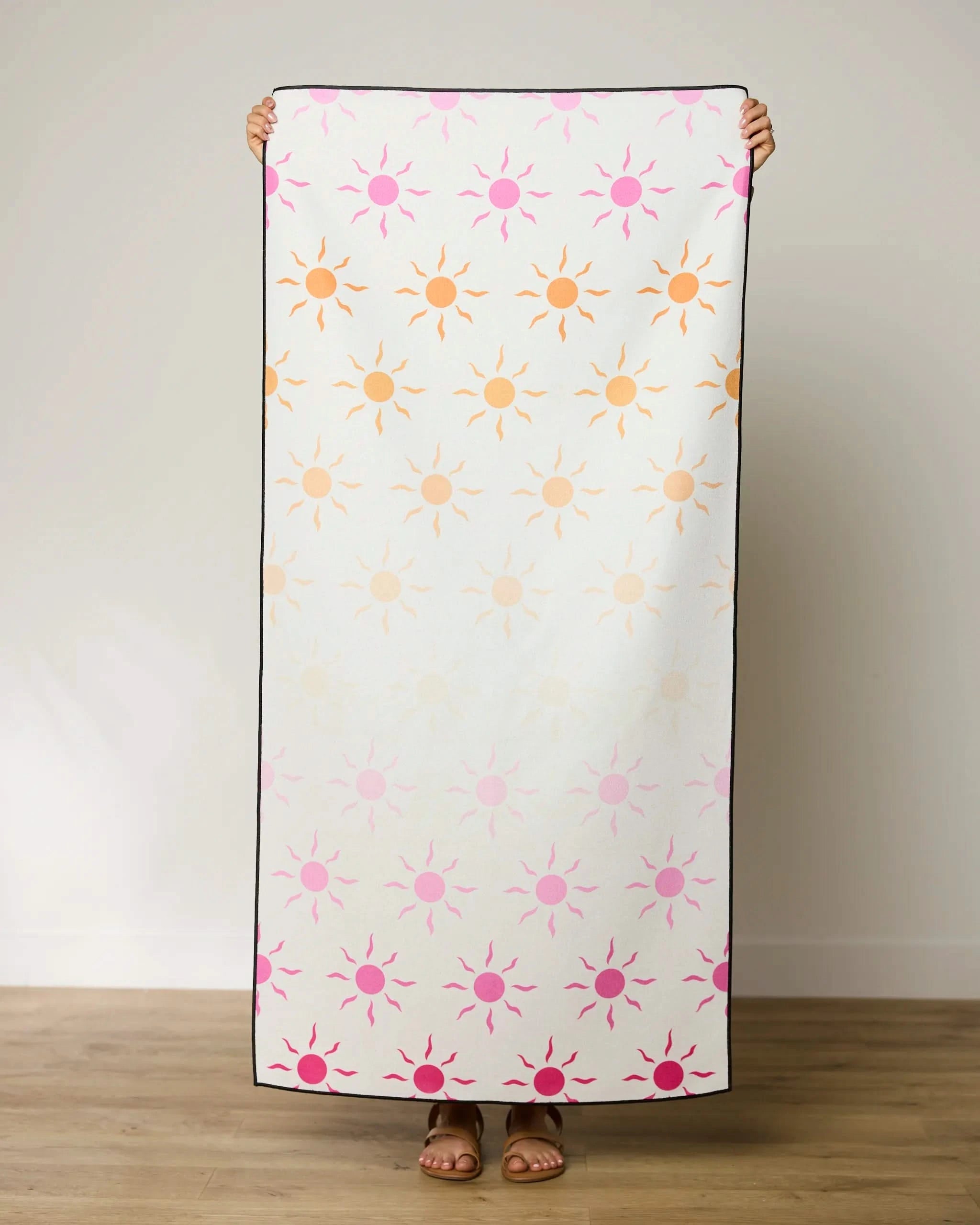 A person stands indoors holding GEOMETRYs Fun in the Sun micro-loop beach towel with orange, pink, and peach sun shapes on white. Only their sandal-clad feet are visible below this quick-drying towel.