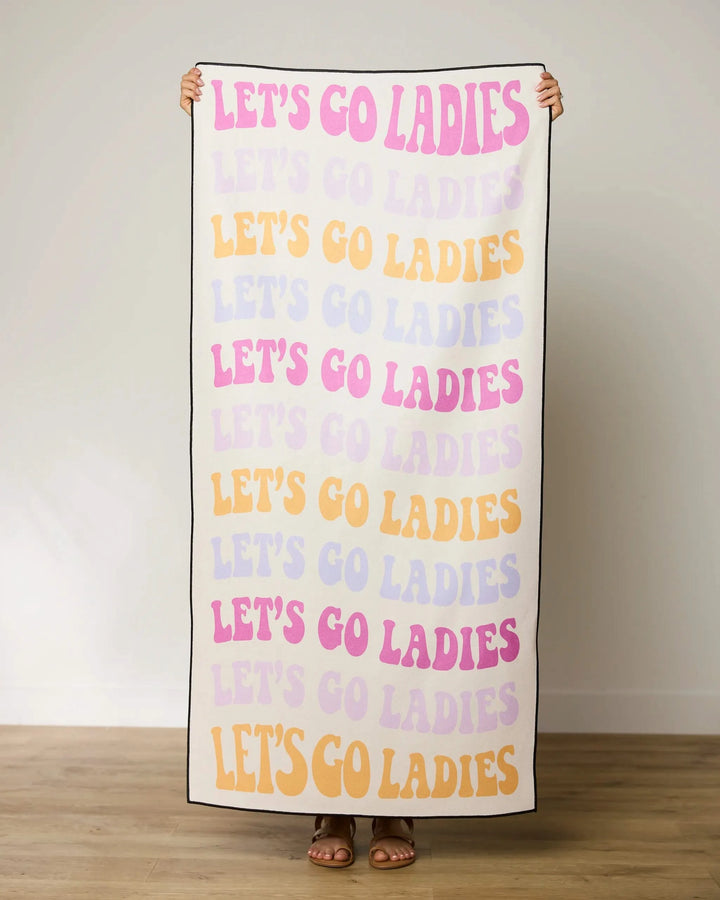 A person stands on a wooden floor holding GEOMETRYs Ladies Only Micro-loop Beach Towel, which features “LET’S GO LADIES” in pink, yellow, and lavender. The quick-drying towel is made from recycled materials; their feet peek beneath it.