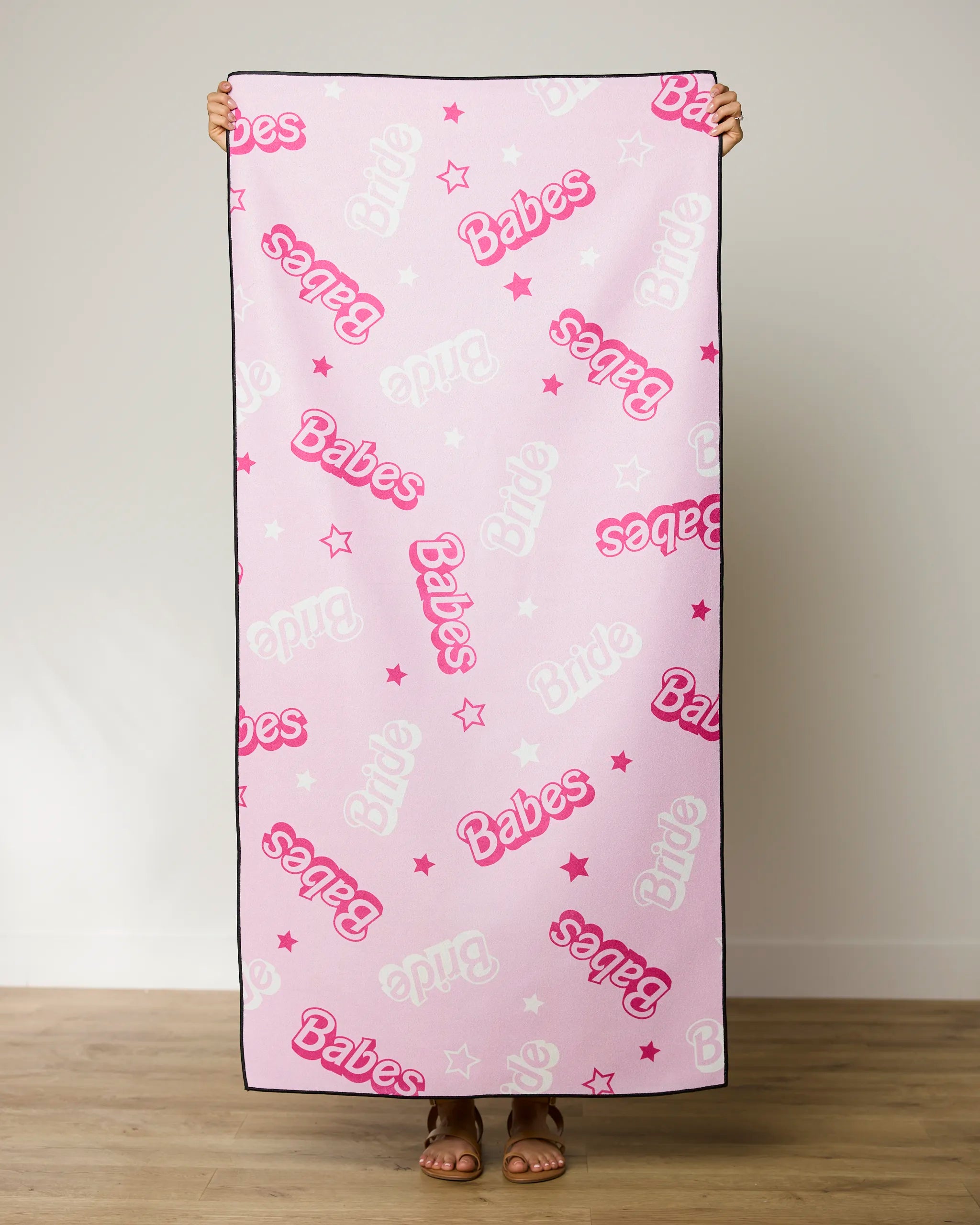 A person holds up GEOMETRY’s Lets Go Party quick-drying beach towel with Babes in bright pink, Bride in white, plus small pink and white stars, against a plain background.
