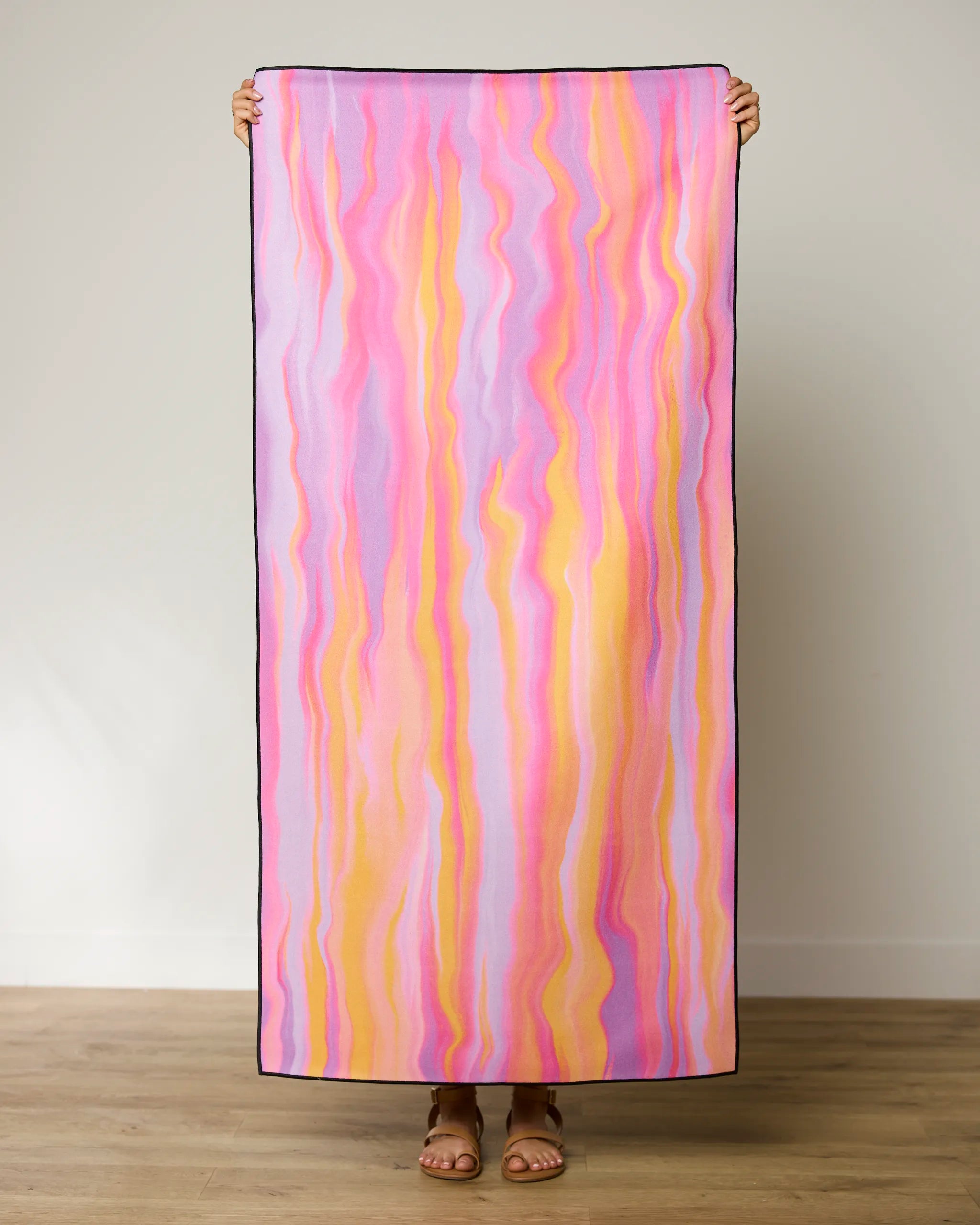 A person holds GEOMETRYs The Rush beach towel, featuring vertical pink, purple, yellow, and orange wavy stripes. Standing by a plain wall and wooden floor, they showcase this quick-drying towel made from recycled materials.