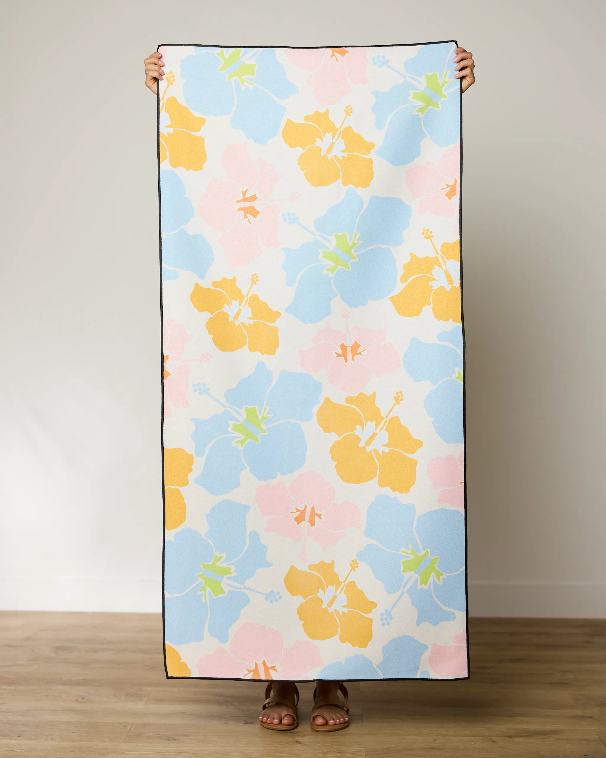 A person holds up the GEOMETRY Hula Hues micro-loop beach towel, featuring vibrant yellow, blue, pink, and green hibiscus flowers. Made from recycled materials, only their feet and hands show against a plain indoor background.