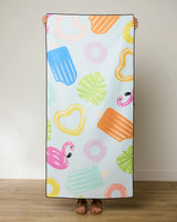 A person displays the GEOMETRY Pool Party quick-drying beach towel, featuring colorful popsicles, flamingos, hearts, donuts, and green leaves on a light background. Only their hands and feet are visible.