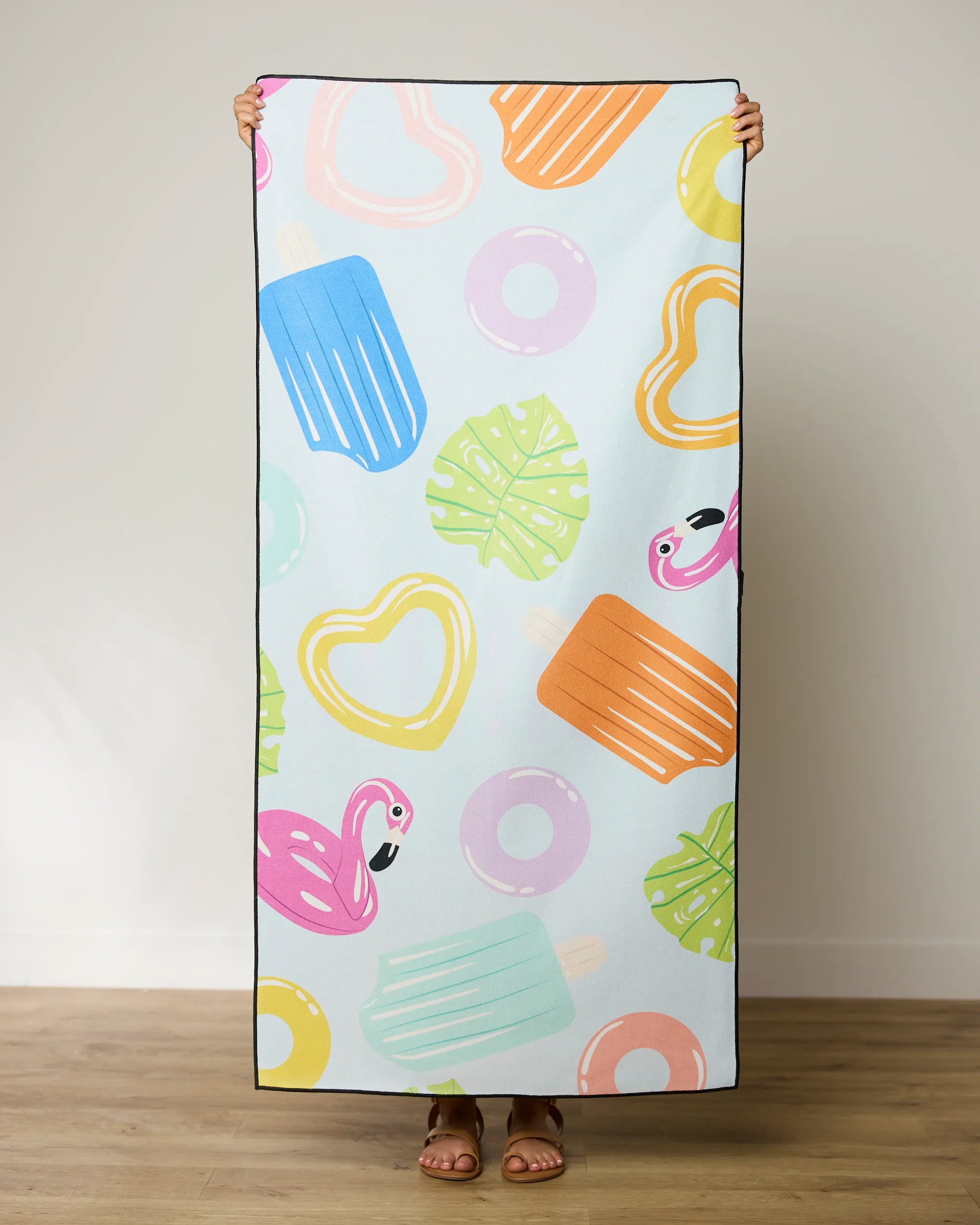 A person displays the GEOMETRY Pool Party quick-drying beach towel, featuring colorful popsicles, flamingos, hearts, donuts, and green leaves on a light background. Only their hands and feet are visible.