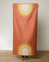 A person stands indoors holding up GEOMETRYs Pumpkin Spice beach towel—a quick-drying, rectangular towel featuring retro rainbow arches in yellow, orange, and beige on a rust background. Only the persons sandal-clad feet are visible.