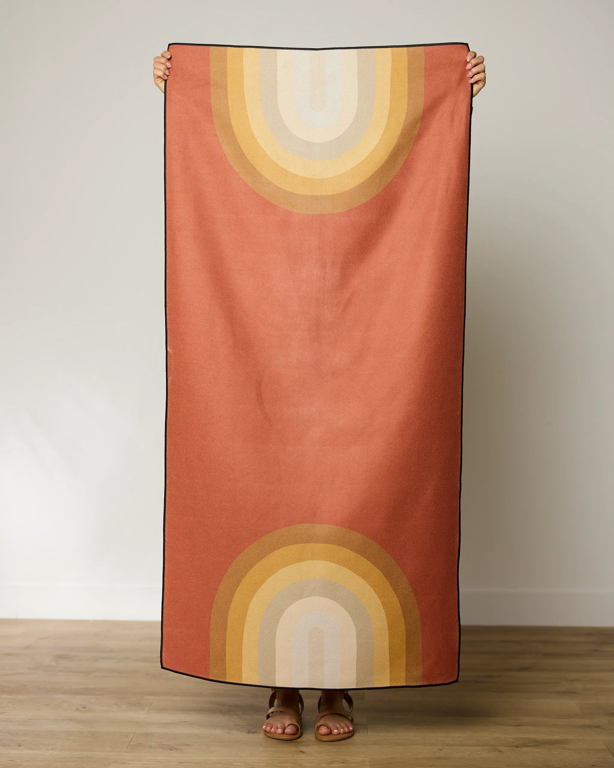A person stands indoors holding up GEOMETRYs Pumpkin Spice beach towel—a quick-drying, rectangular towel featuring retro rainbow arches in yellow, orange, and beige on a rust background. Only the persons sandal-clad feet are visible.