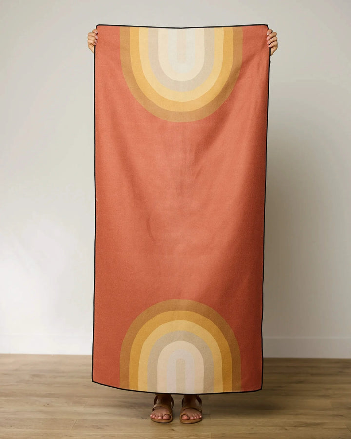 A person stands indoors holding up GEOMETRYs Pumpkin Spice beach towel—a quick-drying, rectangular towel featuring retro rainbow arches in yellow, orange, and beige on a rust background. Only the persons sandal-clad feet are visible.