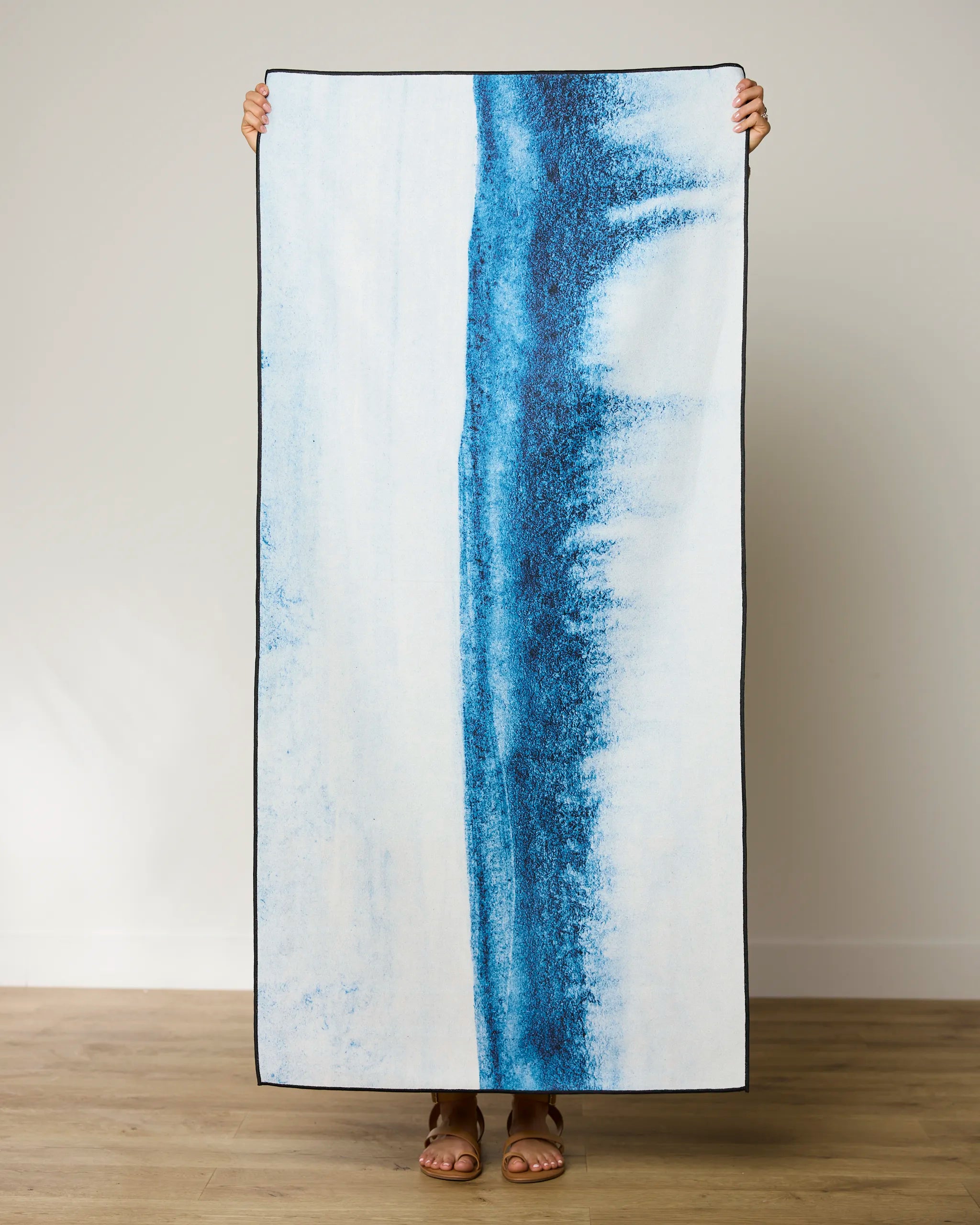 A person stands against a plain wall, holding up the GEOMETRY In The Distance quick-drying beach towel, featuring vertical blue abstract stripes on white. Only their feet and fingers are visible.